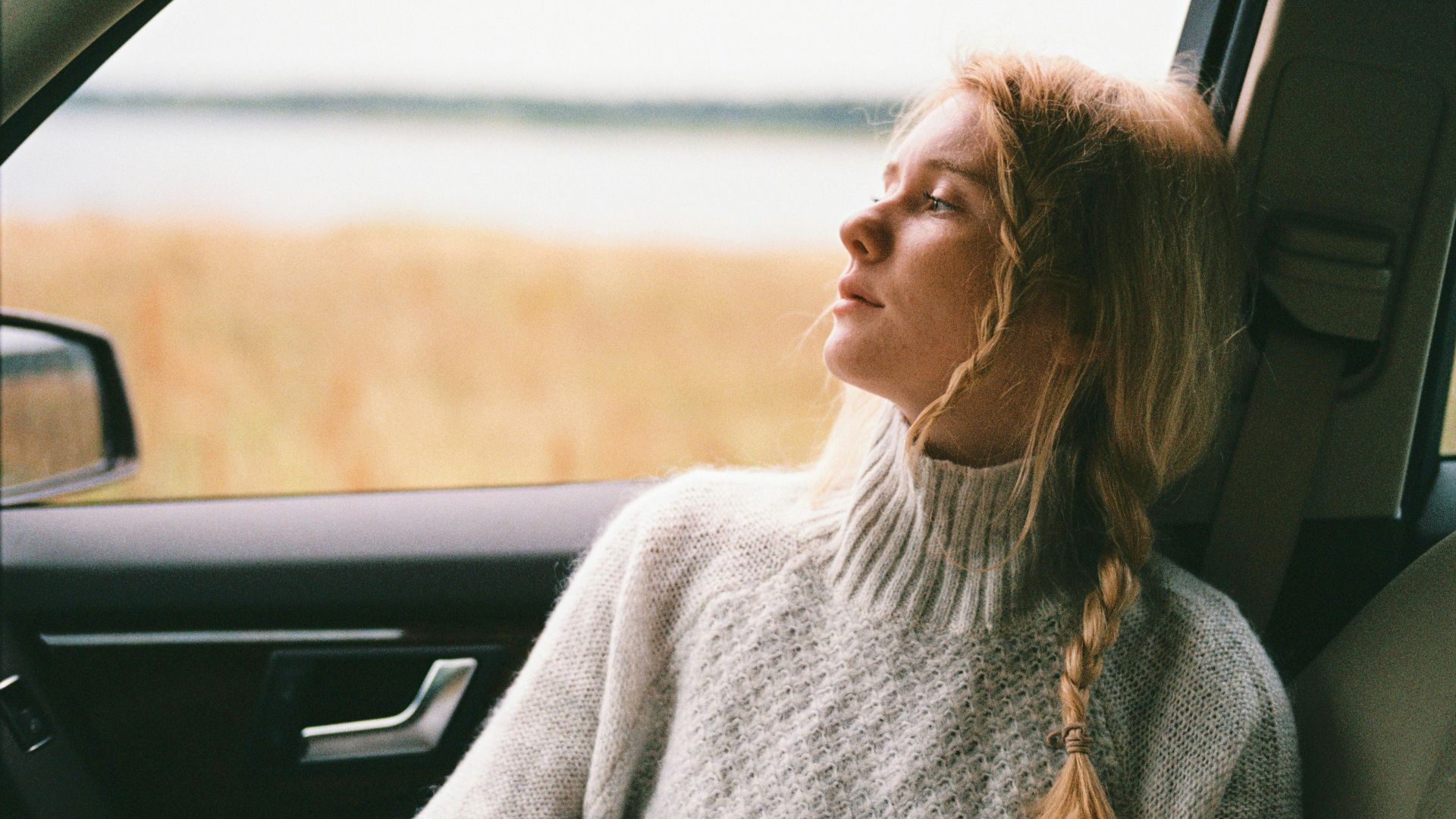 A thoughtful woman with braided hair gazes out a car window during a daytime road trip.