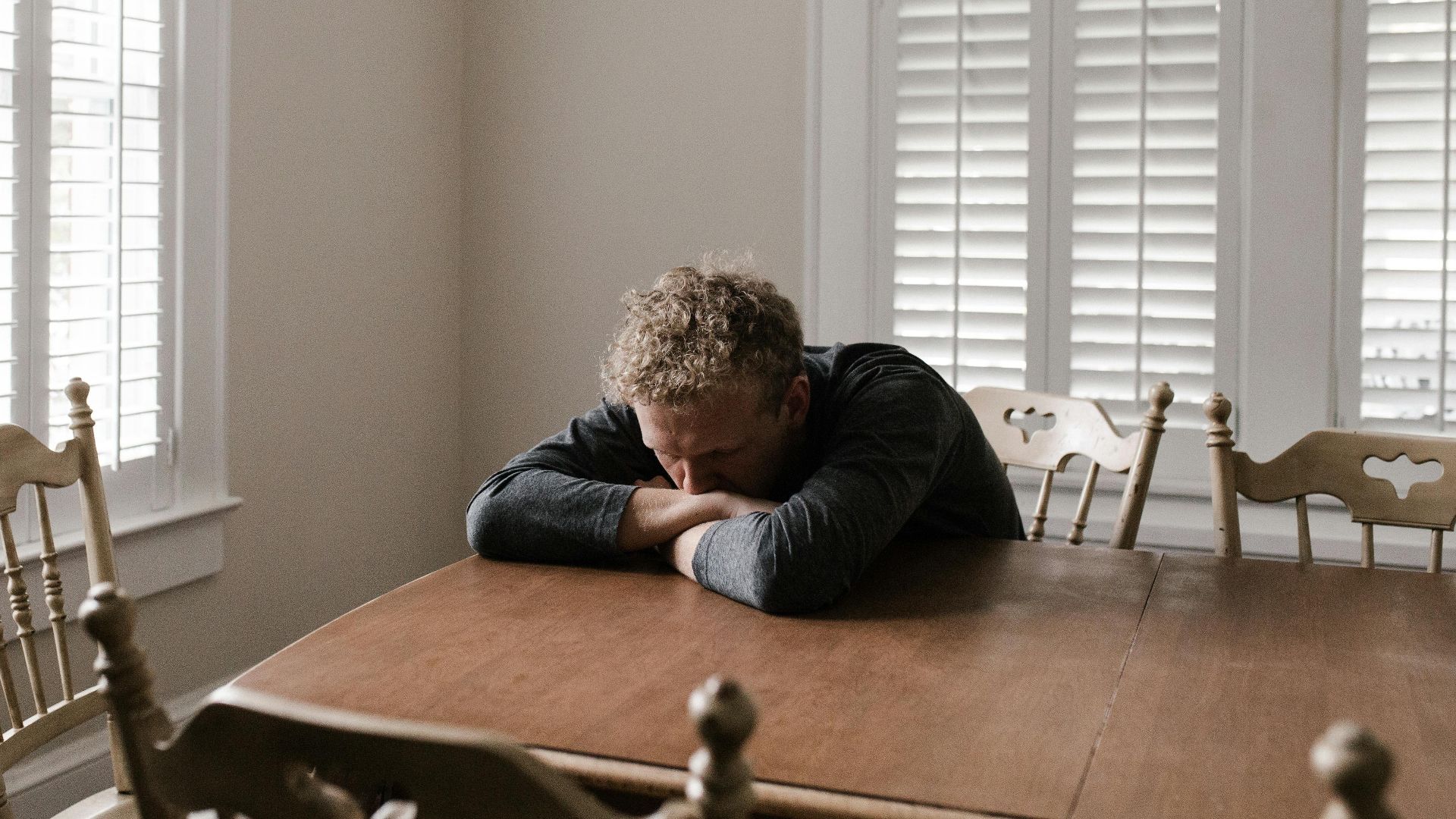 An adult man with emotions of sadness and anxiety sitting alone at a wooden table near windows.