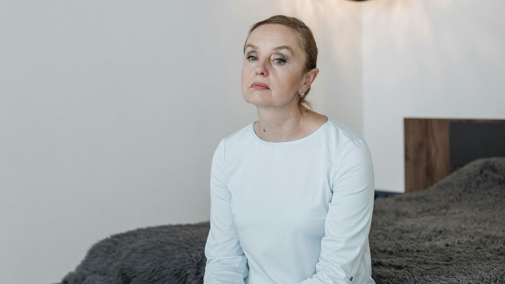Middle-aged woman sitting on a bed in a modern bedroom, looking thoughtful.