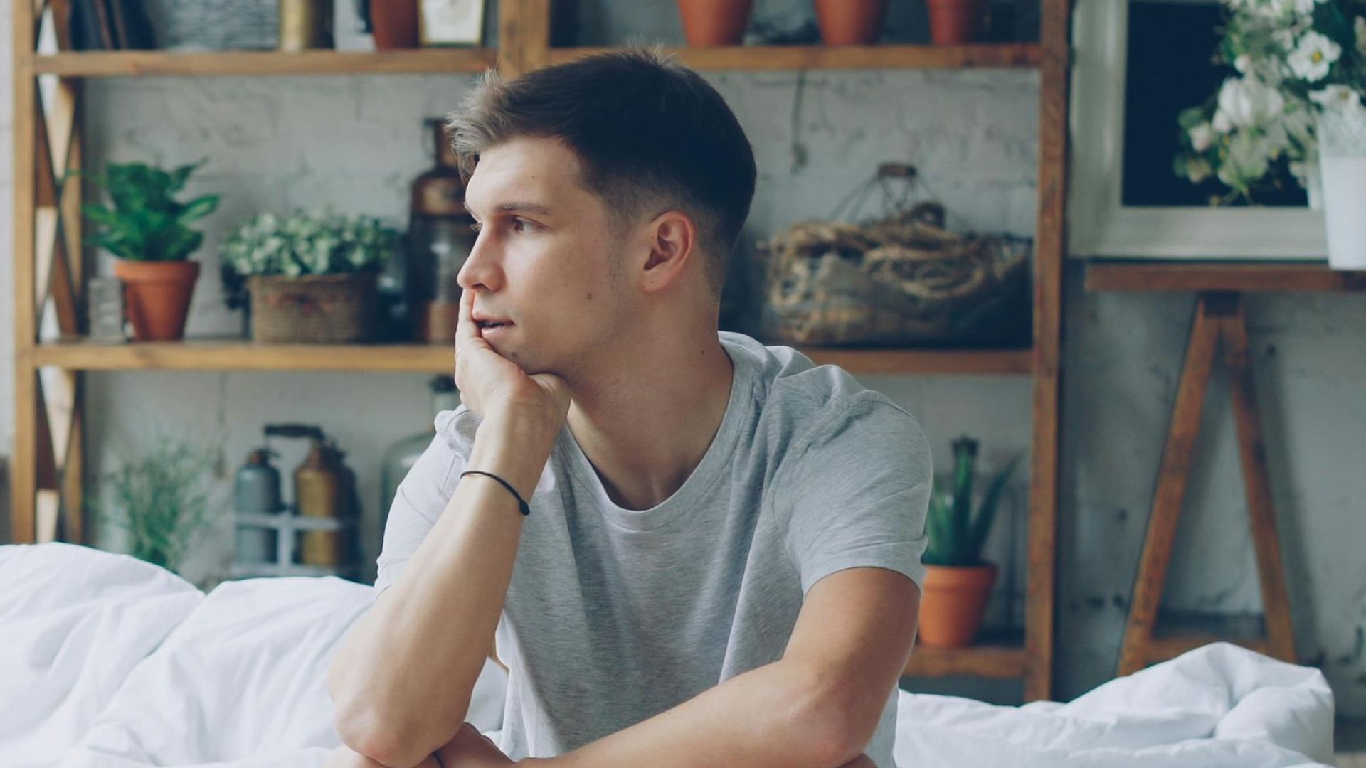 A thoughtful young man sits by a window in a cozy, modern bedroom.