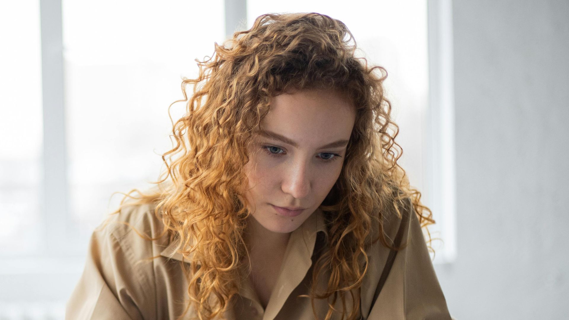 Concentrated female student with curly hair in casual clothes sitting at table and taking notes in notebook while reading book in light room in daytime
