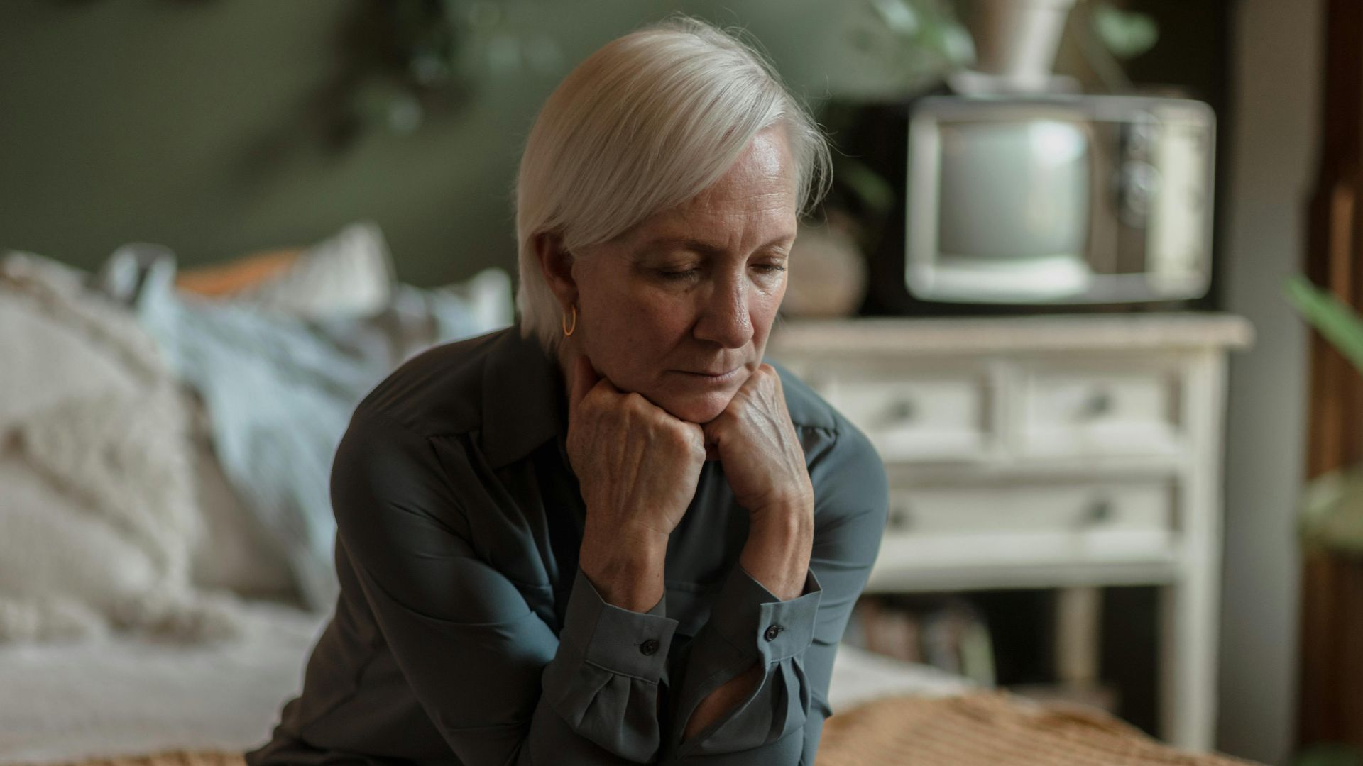 An elderly woman sitting on a bed, deep in thought, with a somber expression in a cozy bedroom.
