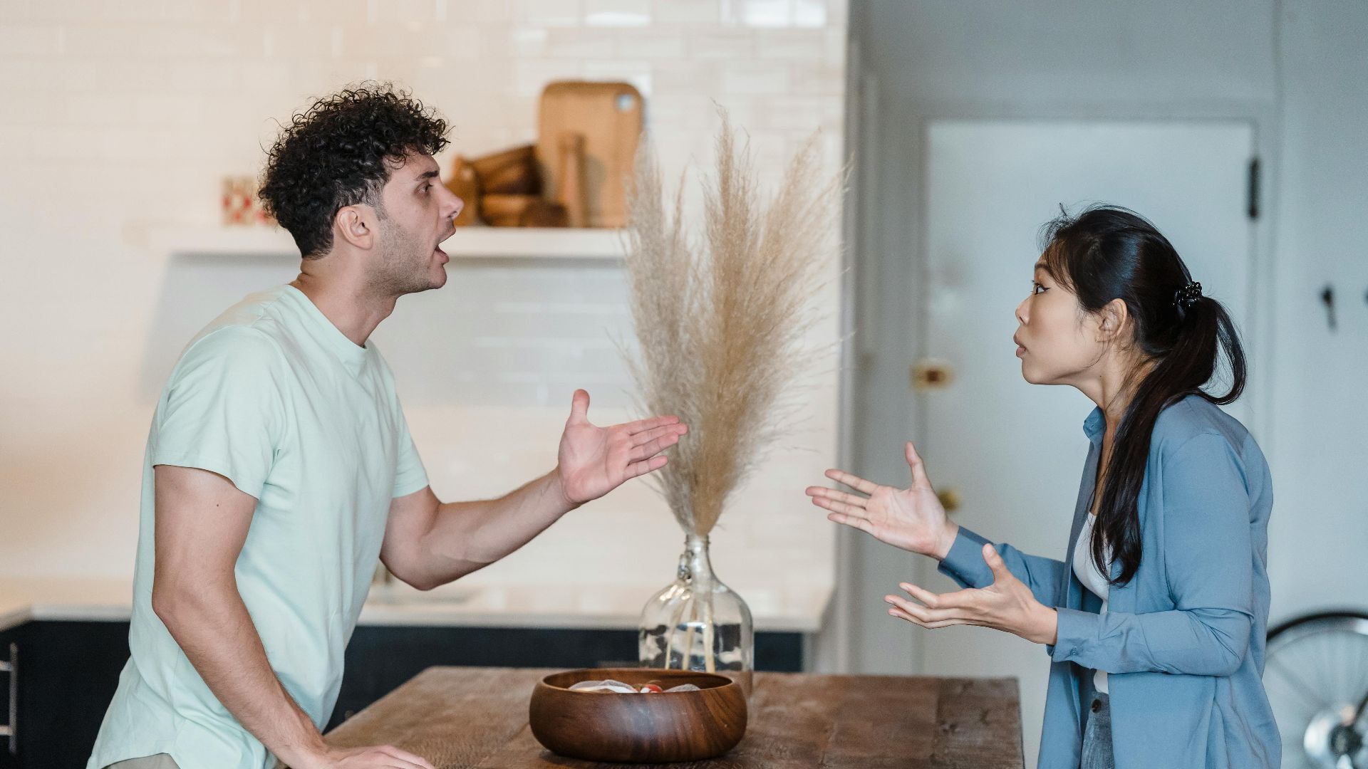 A couple engages in a heated argument at a wooden table in a modern indoor setting.