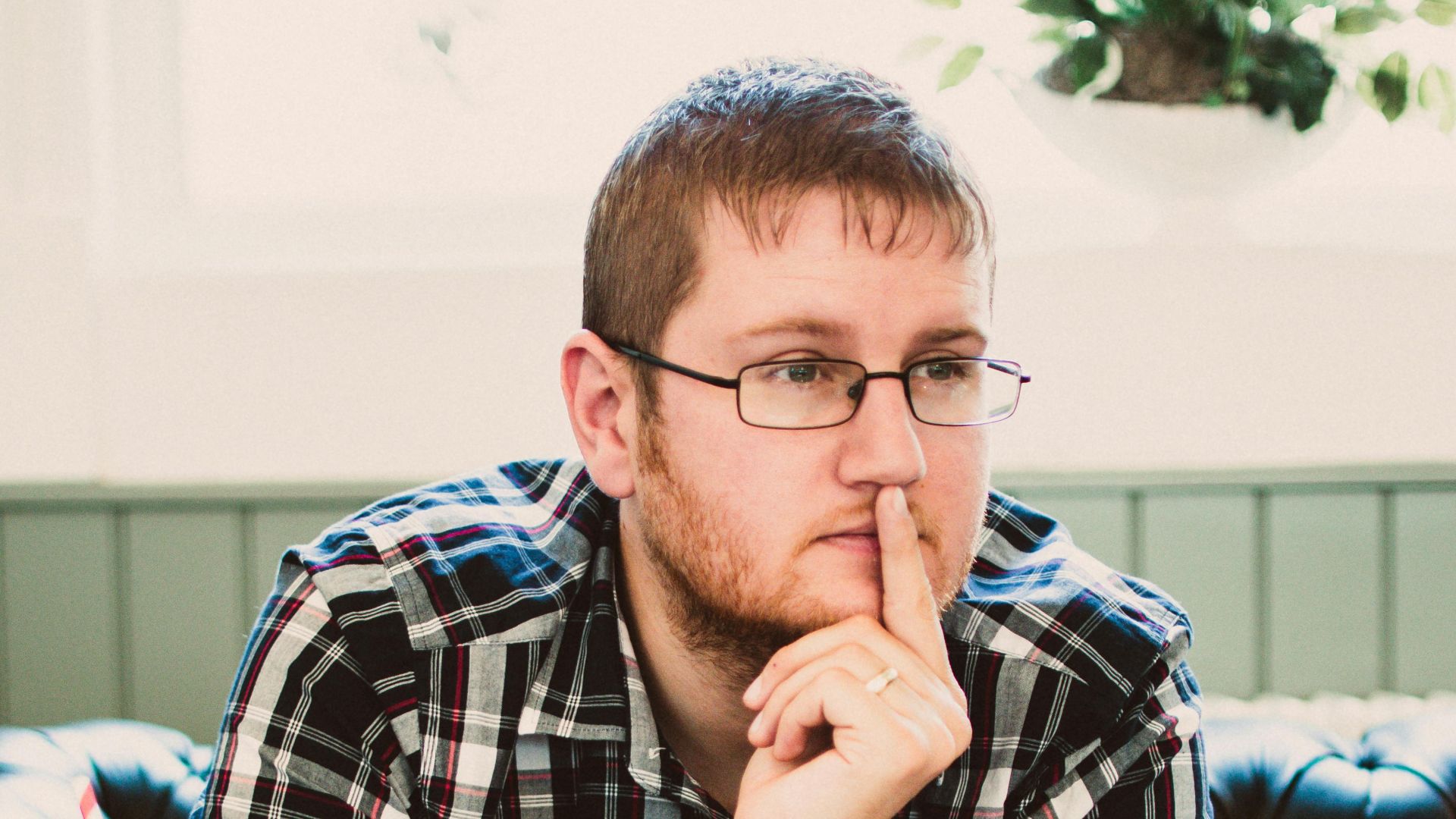 Young man wearing glasses thinking while writing in a cafe with a drink beside him.
