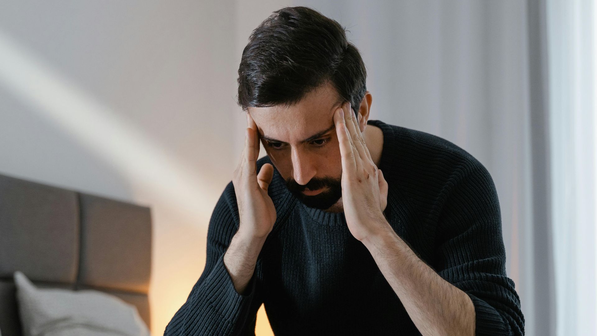 A man in a black sweater sits on a bed, expressing frustration in a bedroom.