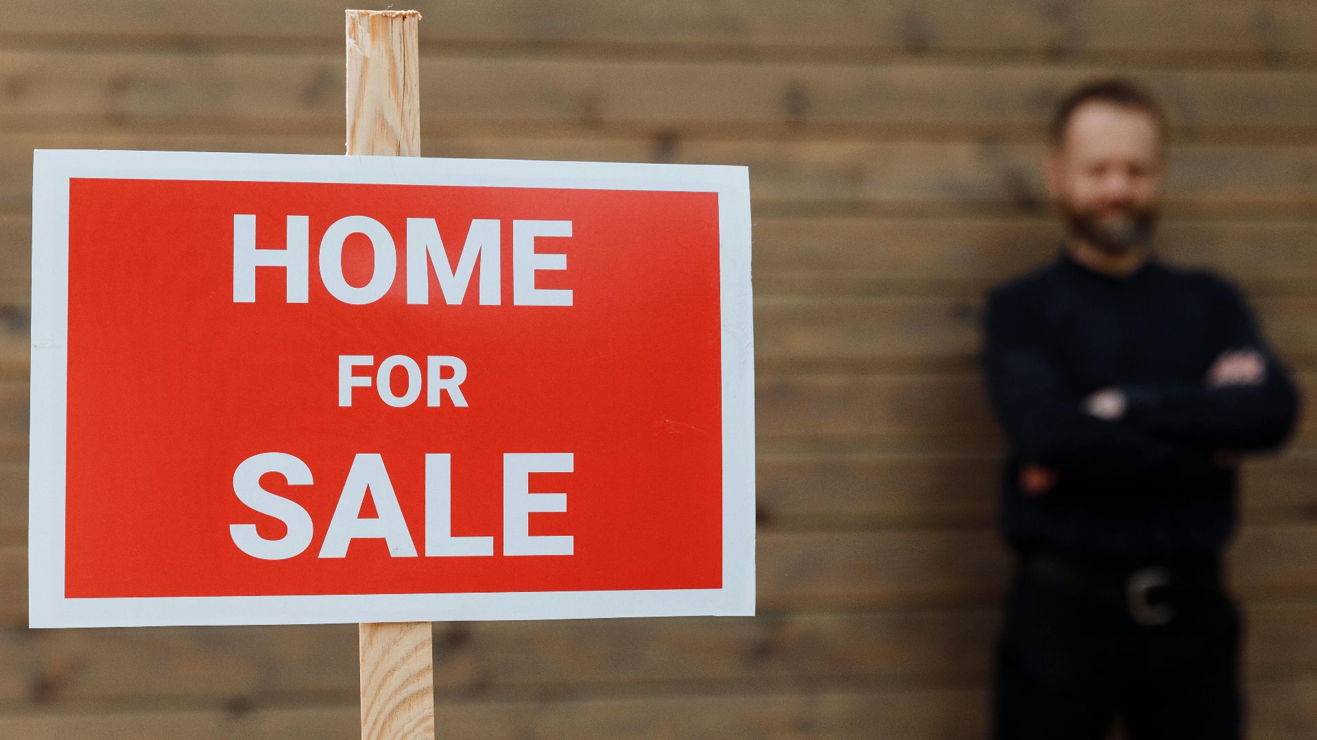A confident real estate agent stands by a home for sale sign against a wooden wall.