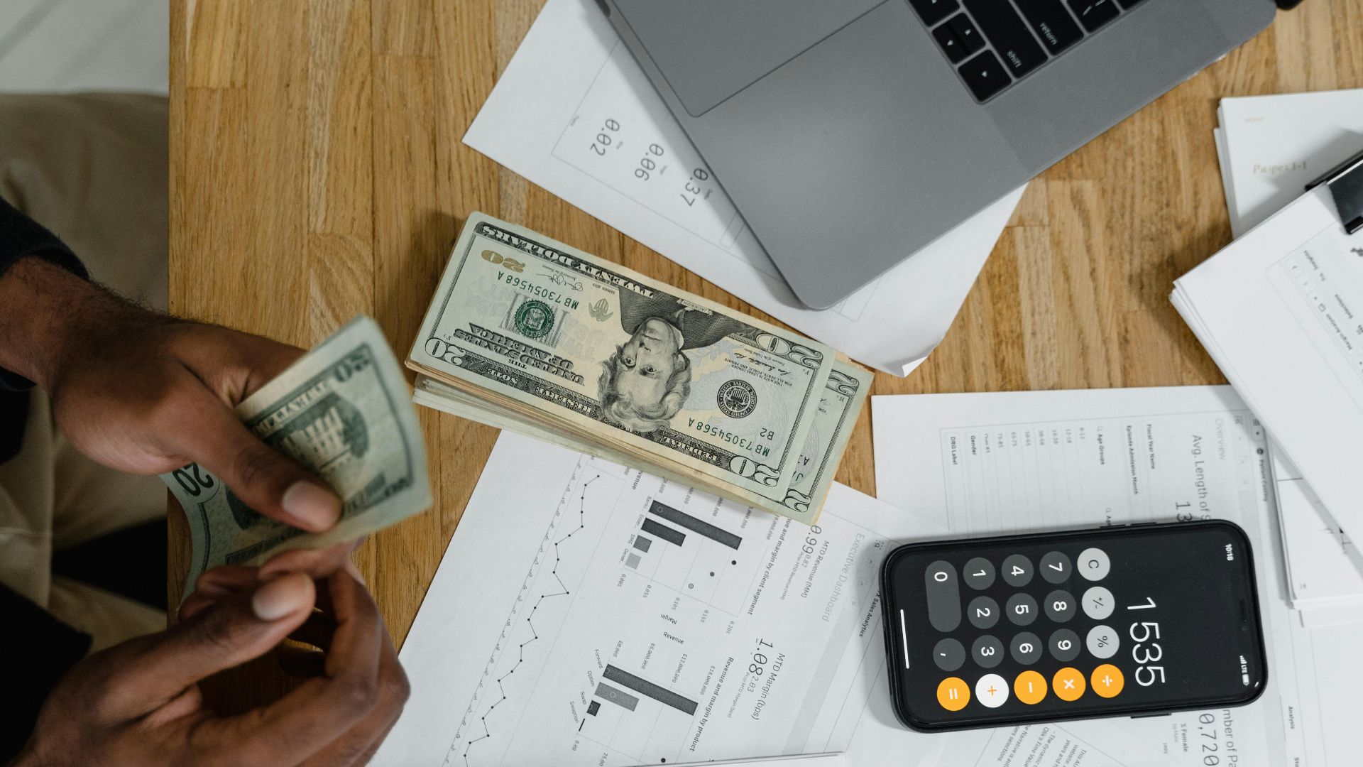 Close-up of hands counting cash on desk with calculator, charts, and laptop, illustrating financial management.