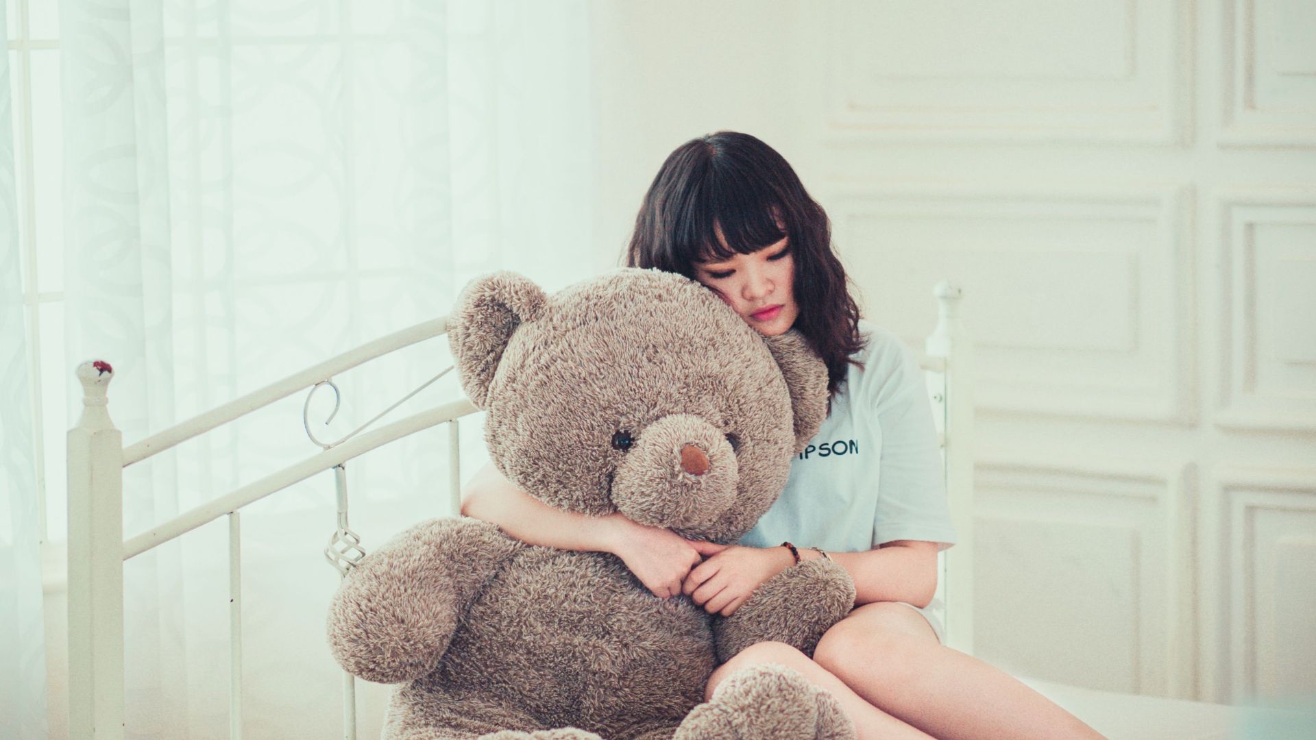 A young woman peacefully hugging a teddy bear while sitting on a bed in a sunlit room.