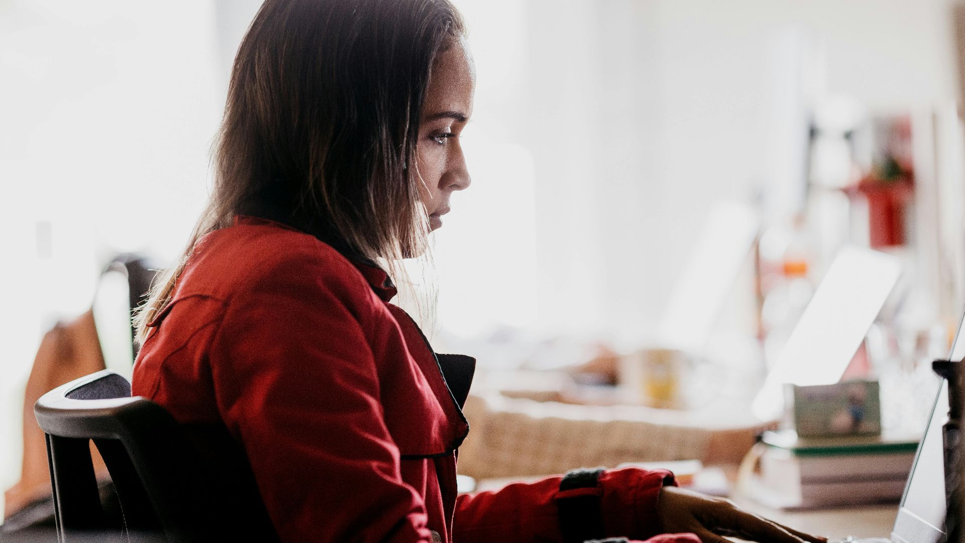 Focused young woman in red jacket typing on laptop in bright workspace.