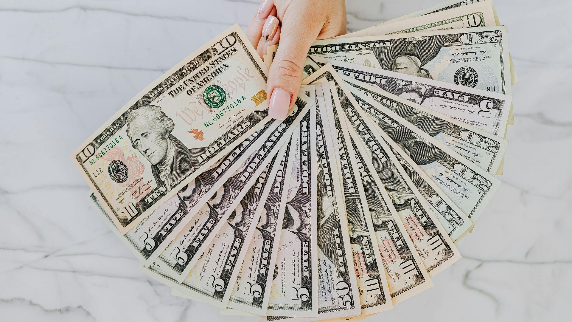 A hand with wristwatch holding a fan of US dollar bills on a marble surface, symbolizing wealth and financial abundance.