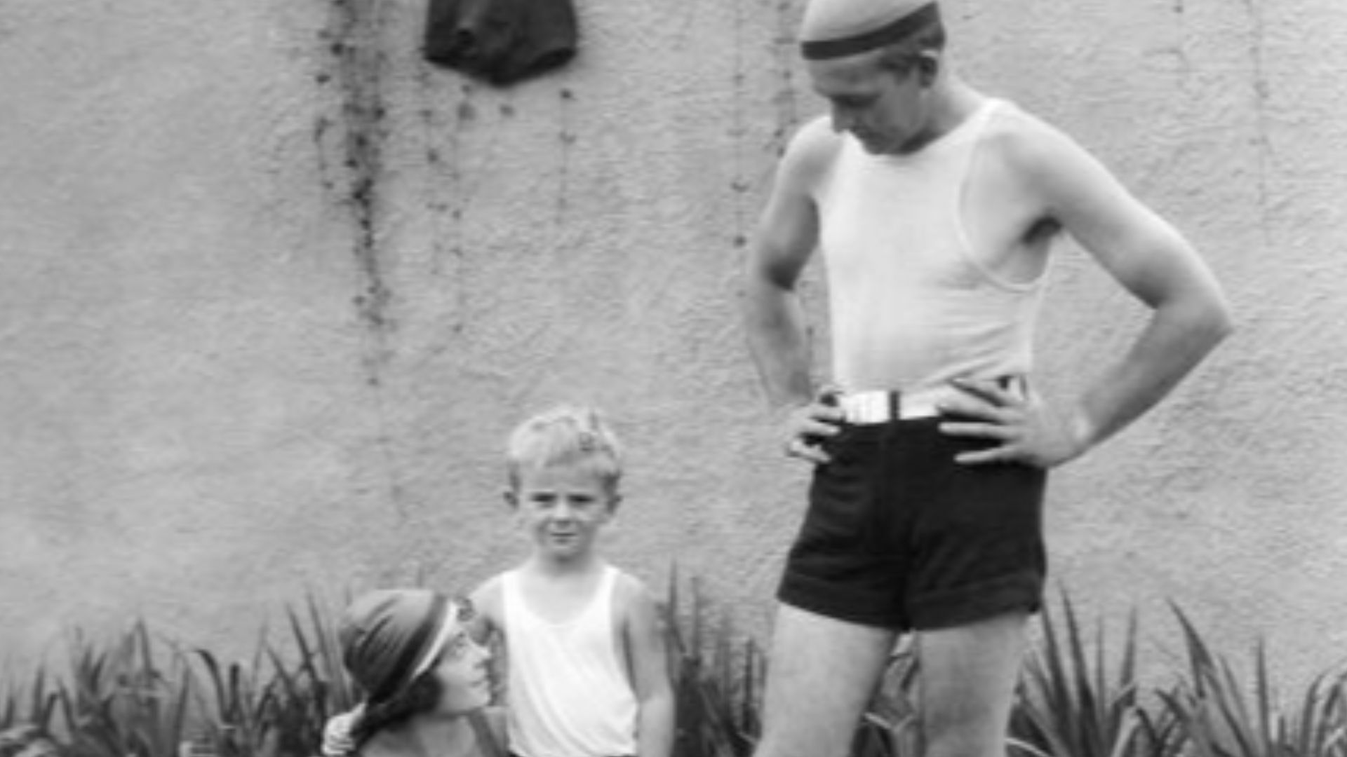 Dorothy Davenport Reid, Wallace Reid, Jr., and the silent film star Wallace Reid in bathing suits at the edge of a swimming pool in a publicity still, c. 1920.