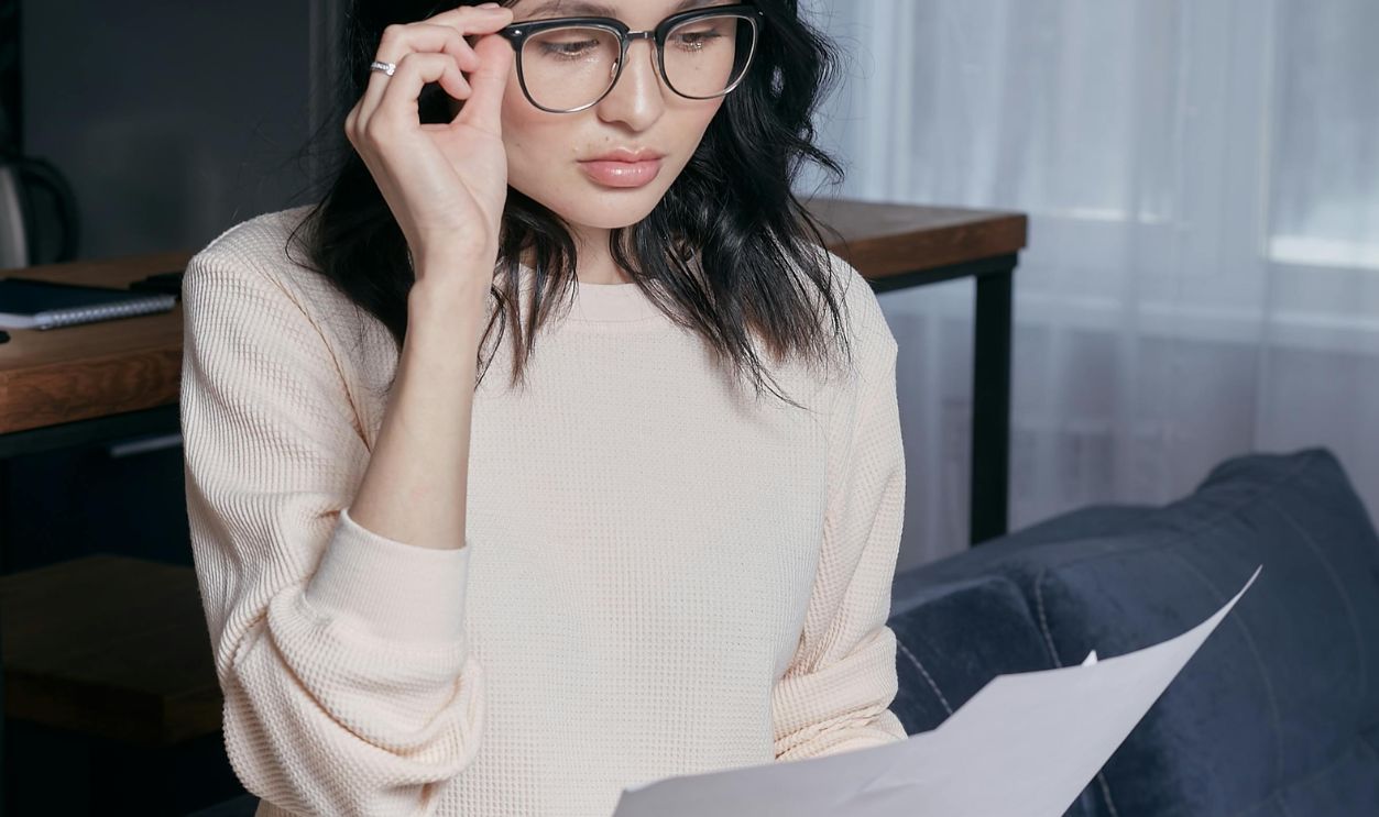 Young woman with eyeglasses assessing financial papers with a concerned look in a modern home setting