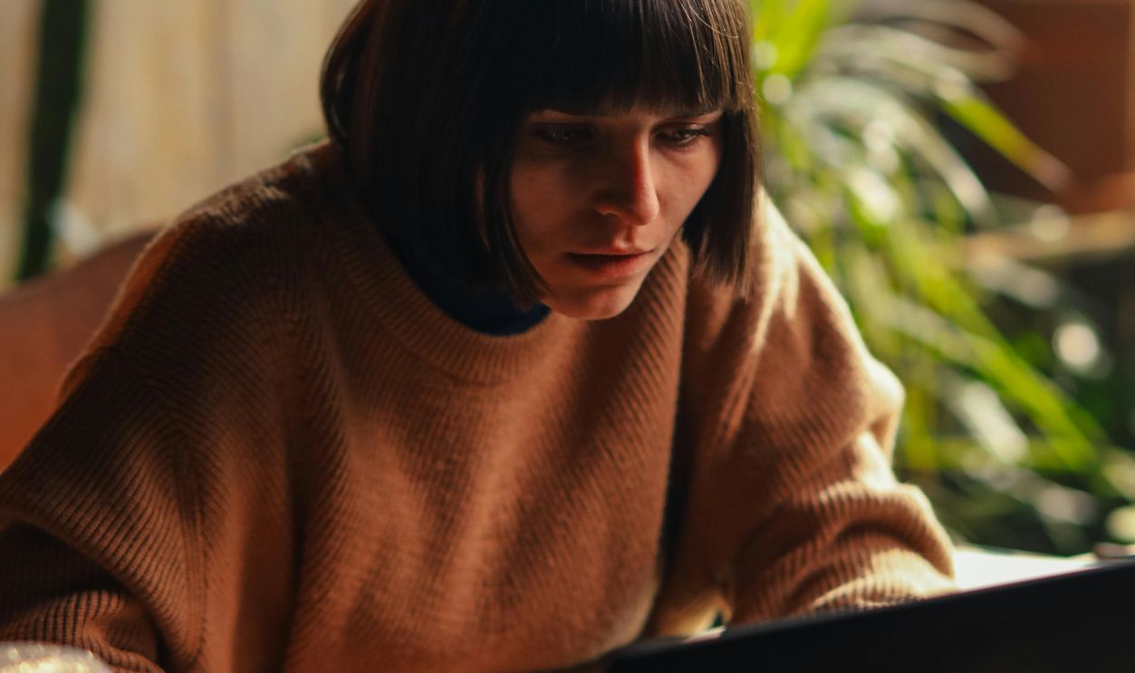 A woman in a cozy sweater concentrates on her laptop in a sunlit room, surrounded by plants
