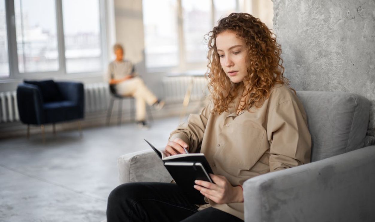 A young woman with curly hair reads a notebook in a bright, spacious loft. Minimalist decor and calm ambiance