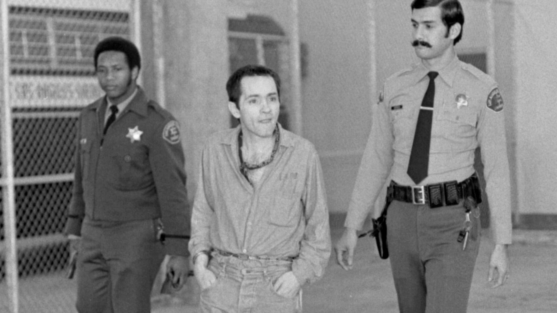 Charles Manson leaving courtroom where he appeared as a witness in behalf of Catherine (Gypsy) Share. Manson, wearing his hair cut short, is manacled hand and foot.