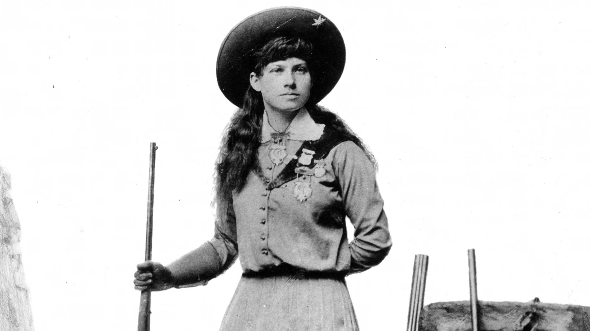 Studio portrait of Annie Oakley. She holds a rifle and poses near two others.