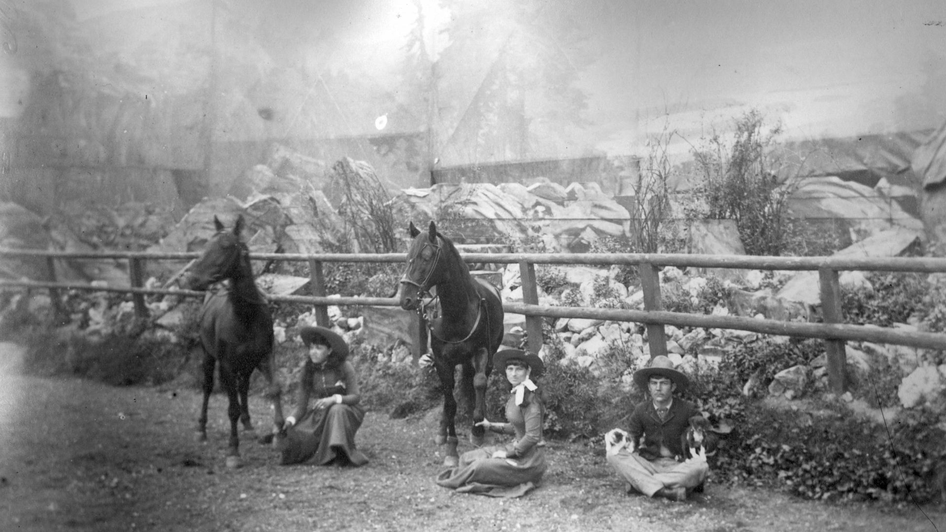 Annie Oakley, center, sits on the ground holding the front hoof of a horse in front of a set for Buffalo Bill's Wild West Show. Another young woman, possibly Della Ferrell, sits near her, also in front of a horse. A young man sits on the ground in the rig