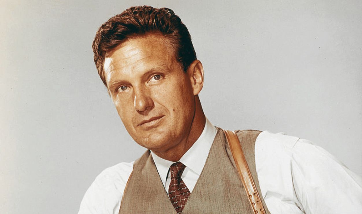 Robert Stack (1919-2003), US actor, wearing a grey waistcoat with a white shirt and a dark red tie, with a tan leather holster over his shoulder in a studio portrait, against a light blue background, issued as publicity for the US television series, 'The Untouchables', USA, circa 1961. The police drama starred Stack as 'Eliot Ness'. 