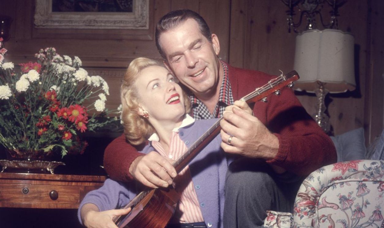 circa 1955: American actor Fred MacMurray shows his wife, actor June Haver, how to play a guitar while sitting behind her on the floor of a living room. 