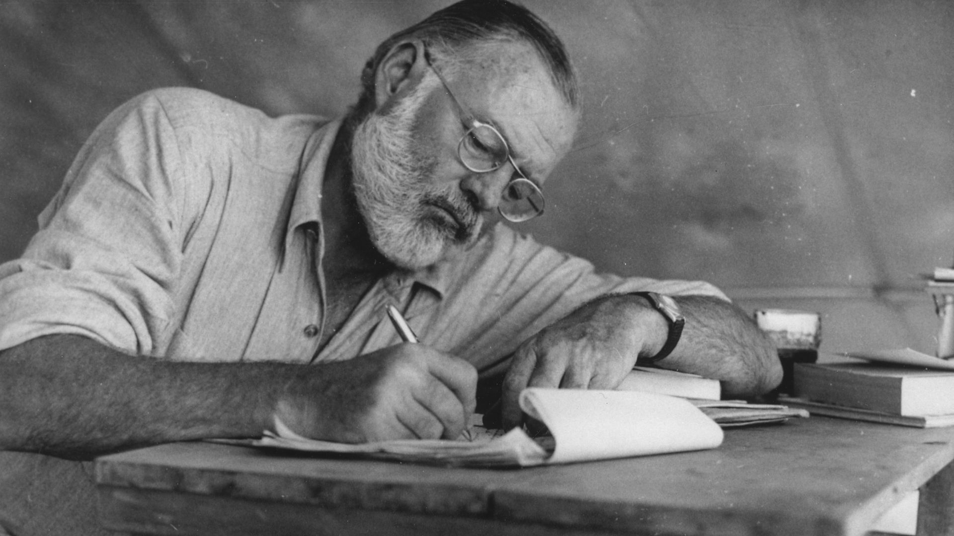 Scope and content:  Photograph of Ernest Hemingway sitting at a table writing while at his campsite in Kenya.