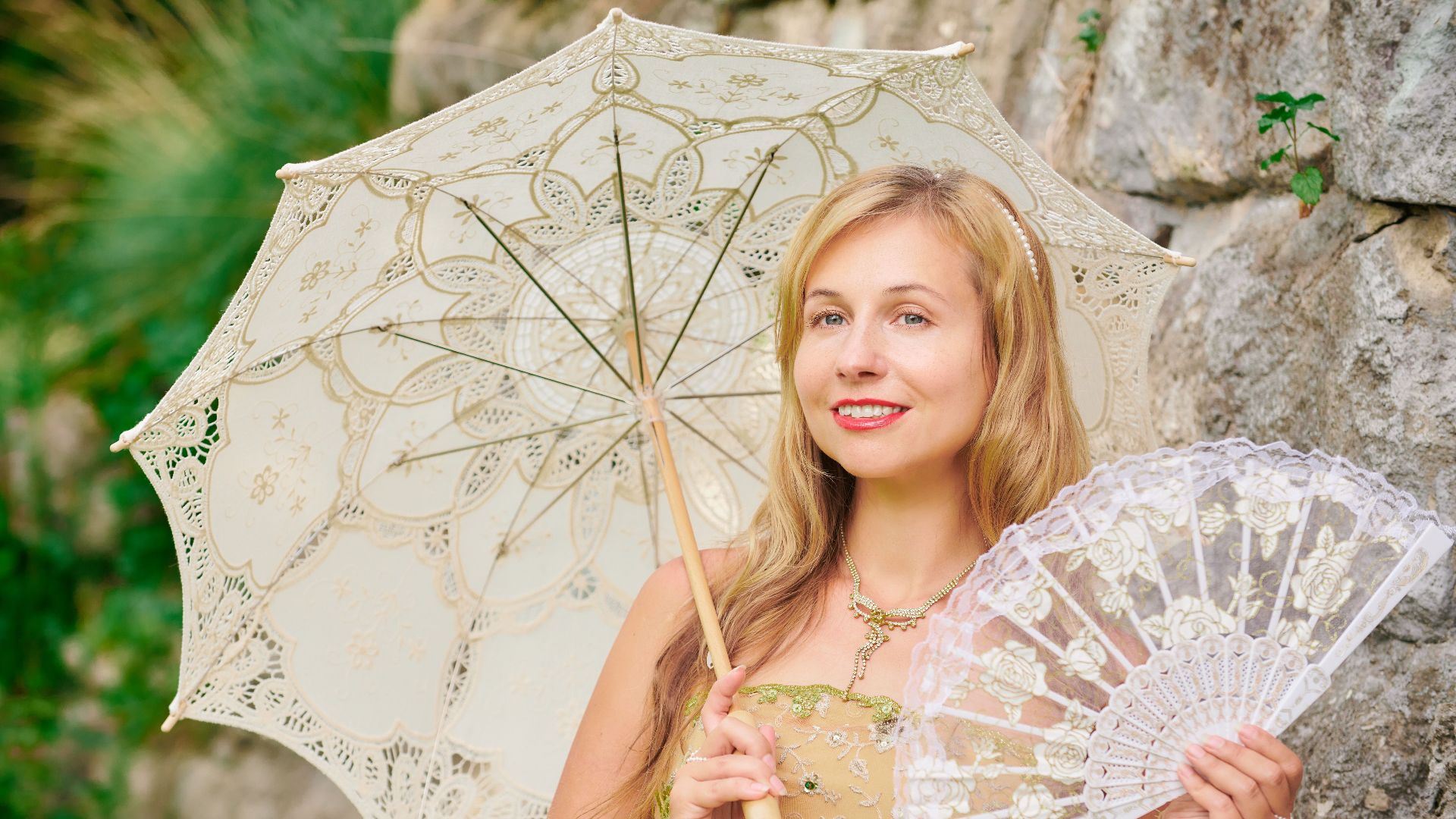 Portrait of a woman in vintage attire with a lace umbrella and fan in a garden setting.