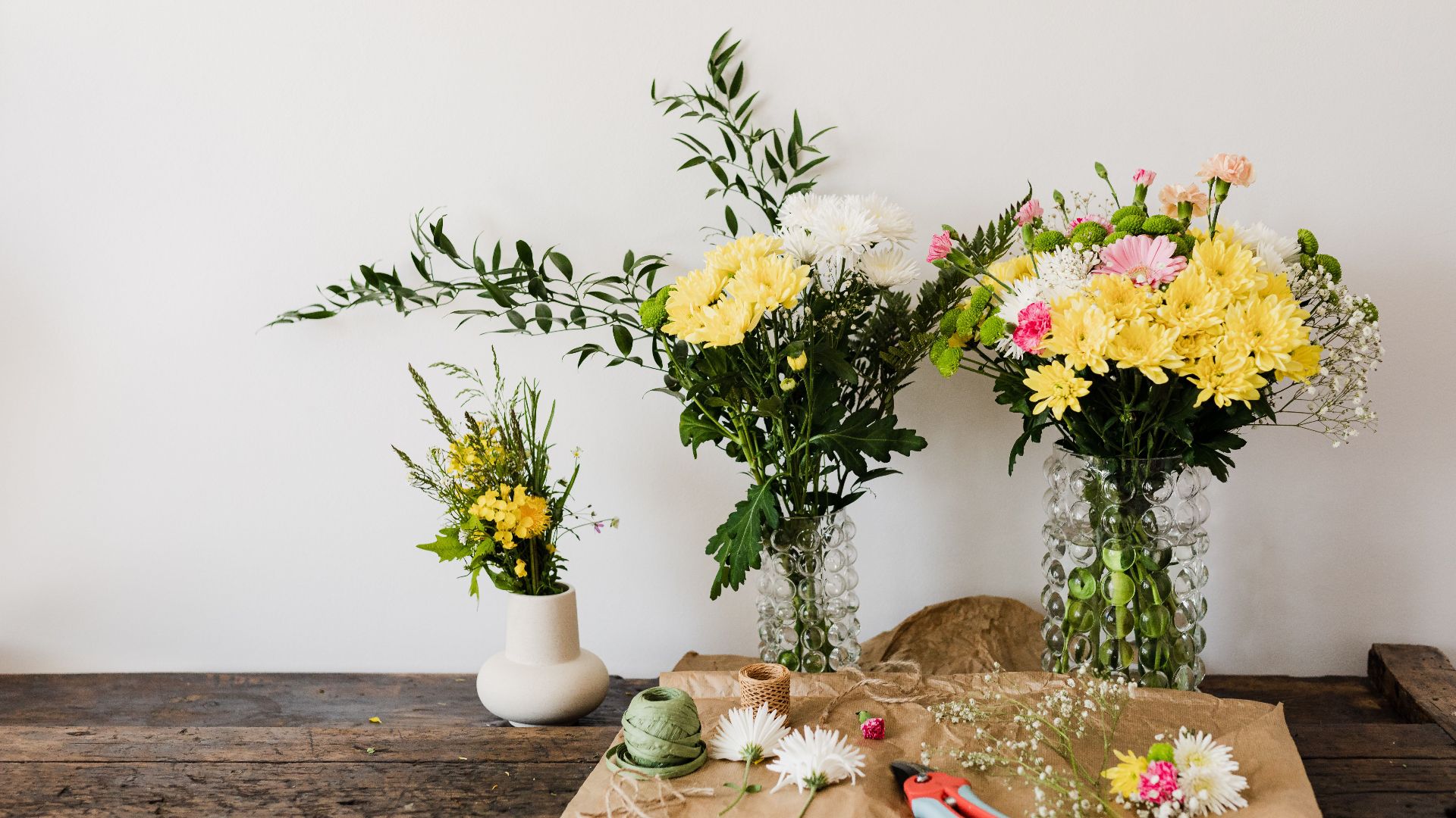 Exuberant mum flower bouquets in glass vases on rustic dark brown table with pruner and twine on craft paper beside