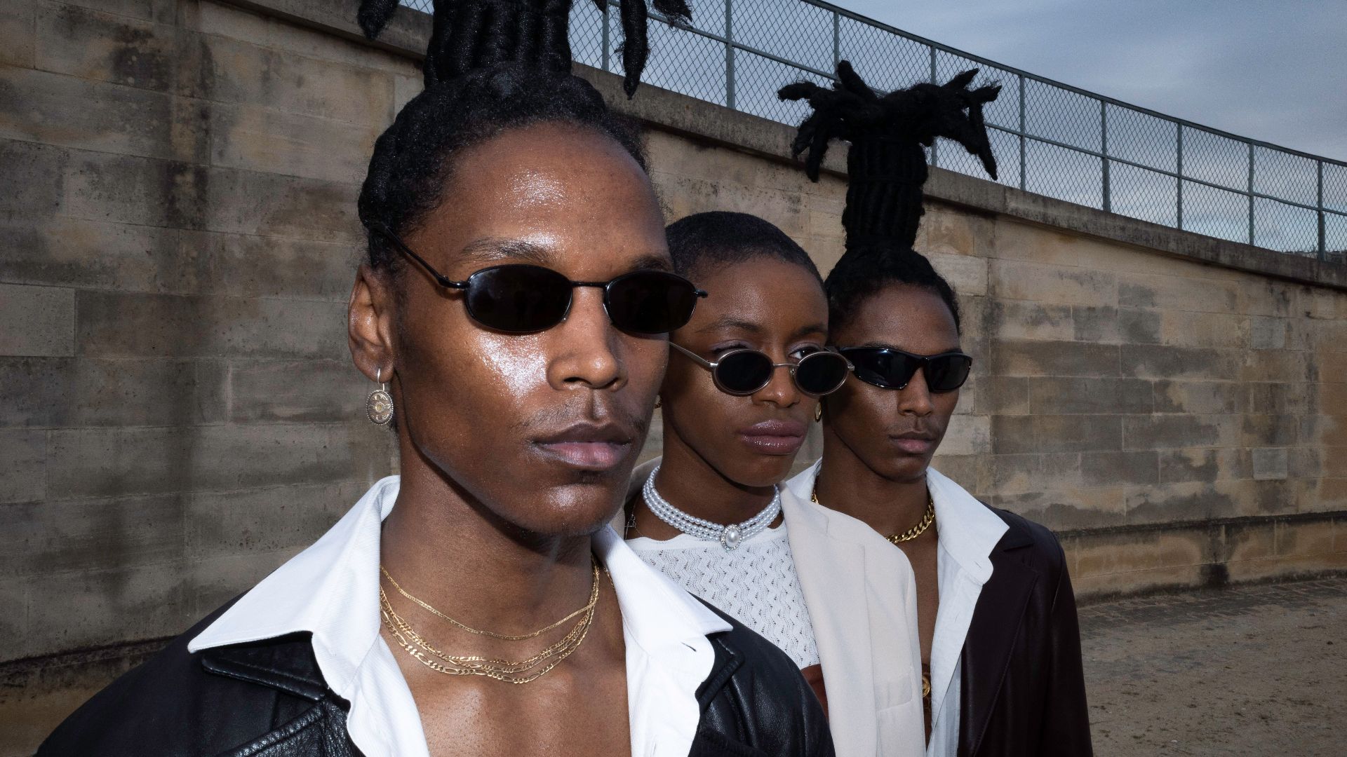 A stylish trio of black adults wearing sunglasses with unique hairstyles, posing outdoors.