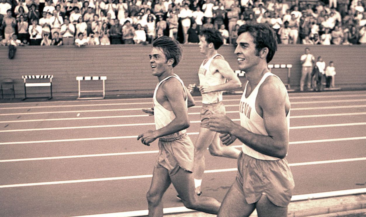 EUGENE, OREGON - JUNE 1970: University of Oregon distance runners Steve Prefontaine (L), Roscoe Divine (R), and a teammate run in front of the grandstands surrounding a race at the university's Hayward Field in Eugene, Oregon in June of 1970. Prefontaine later set the American record for the 5,000 meters at the 1972 Olympic Trials and competed in the 1972 Olympic Games in Munich, Germany. He died in a tragic single-car auto accident in 1975.