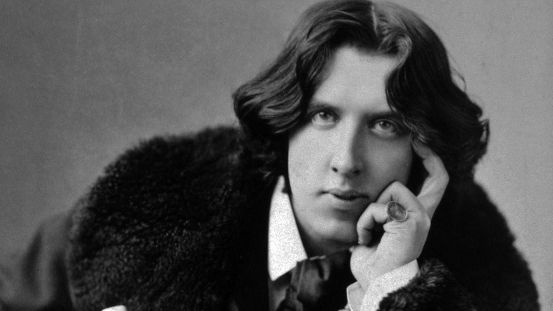 Oscar Wilde, three-quarter length portrait, facing front, seated, leaning forward, left elbow resting on knee, hand to chin, holding walking stick in right hand, wearing coat.