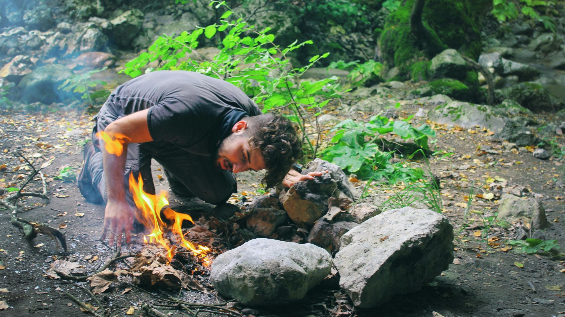 Man attempts to start a campfire surrounded by rocks in a lush green forest.