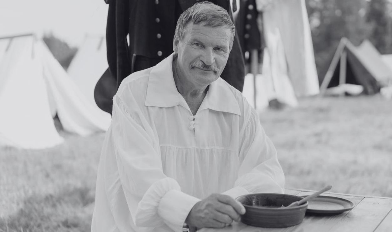 A man in historical attire enjoying a meal at a reenactment camp, outdoors