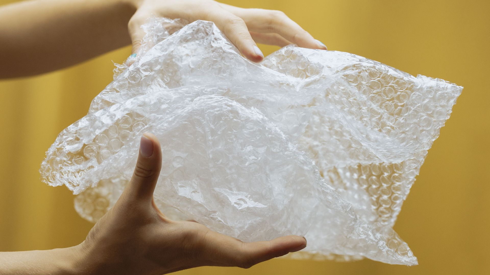 Close-up of hands holding and crumpling bubble wrap against a yellow backdrop indoors.