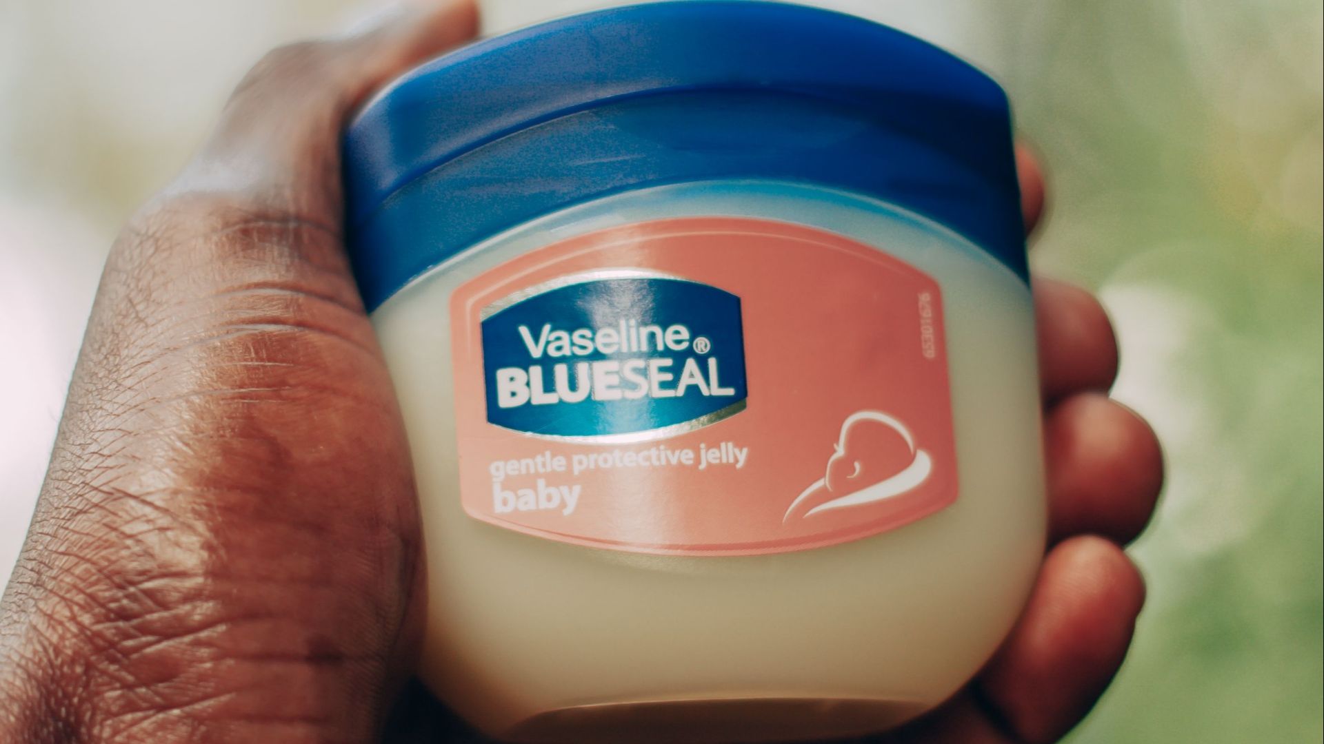 A hand holding a jar of Vaseline Blue Seal gentle protective jelly against a blurred background.