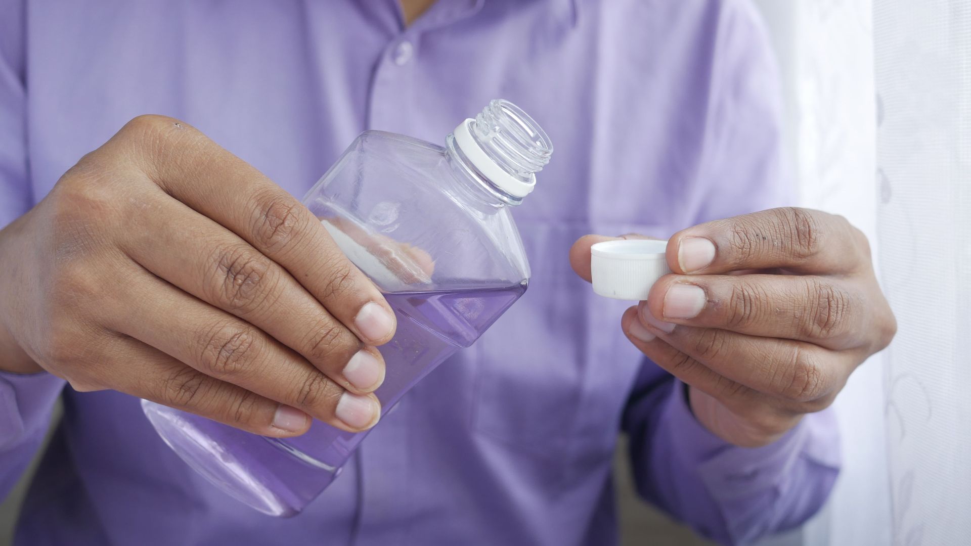 Close-up of hands holding a purple plastic bottle while pouring mouthwash into a cap.