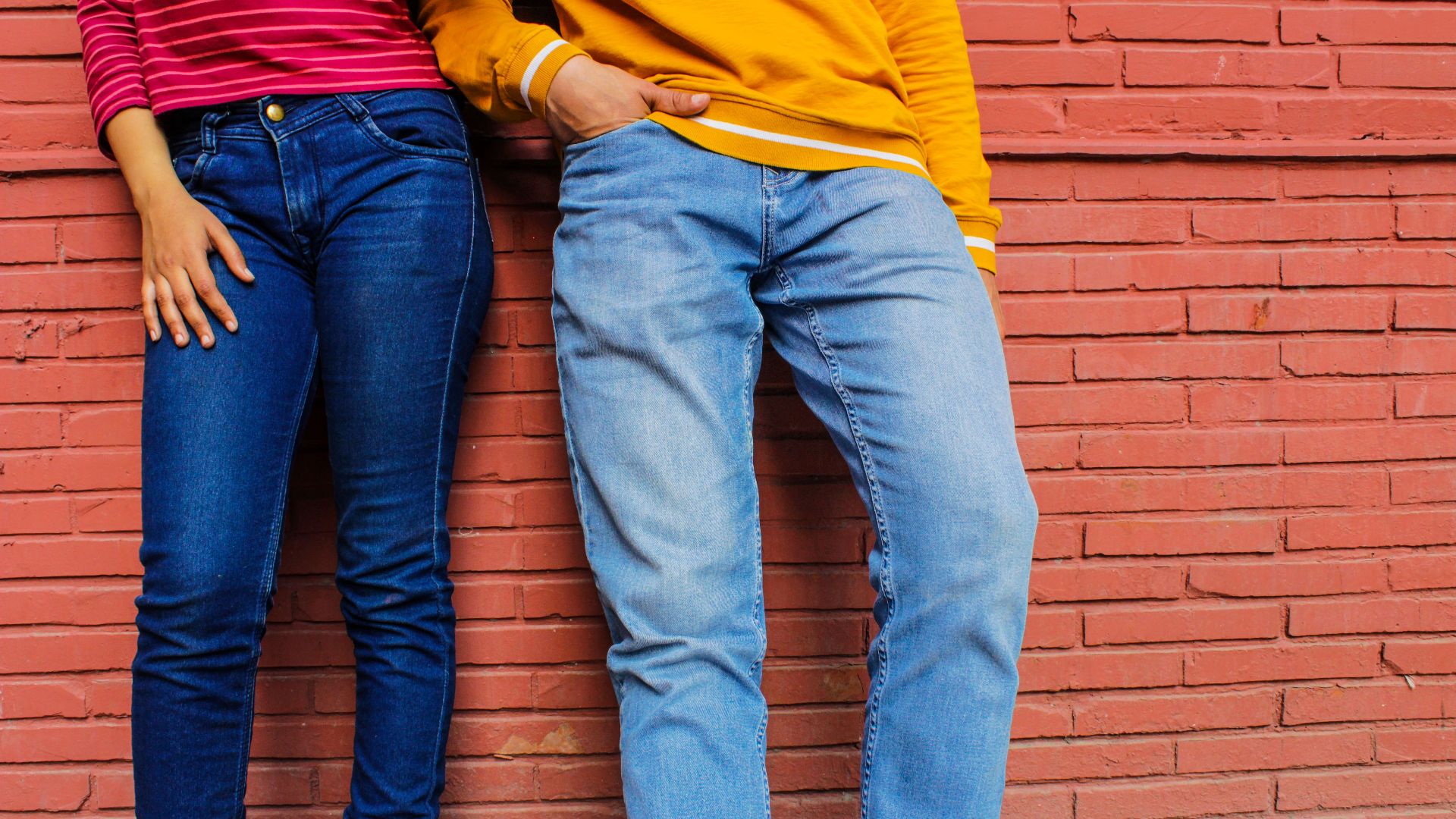 Two people in casual attire and denim jeans against a brick wall.
