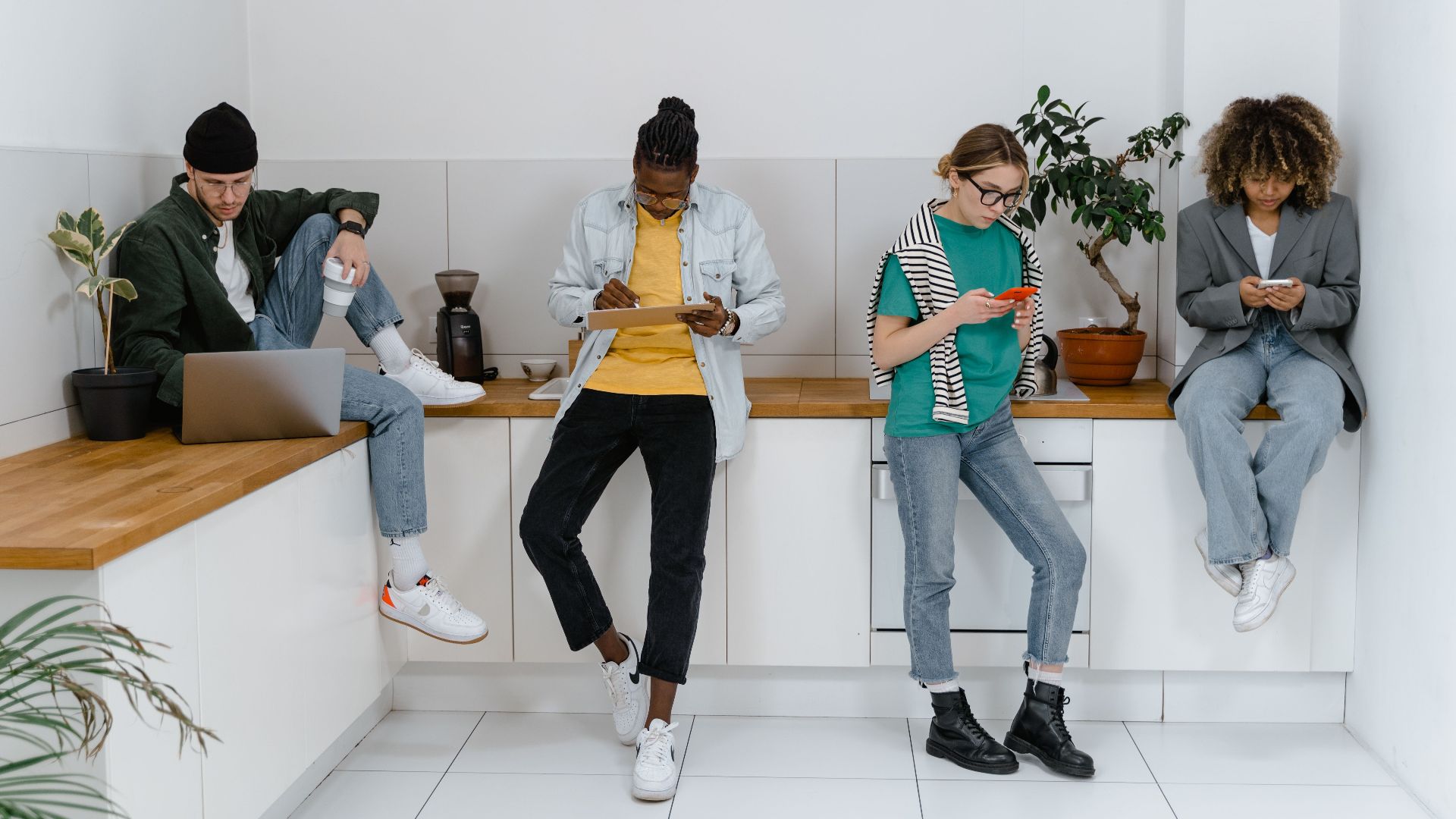 Young adults on digital devices in a modern office kitchen area, emphasizing diversity and teamwork.