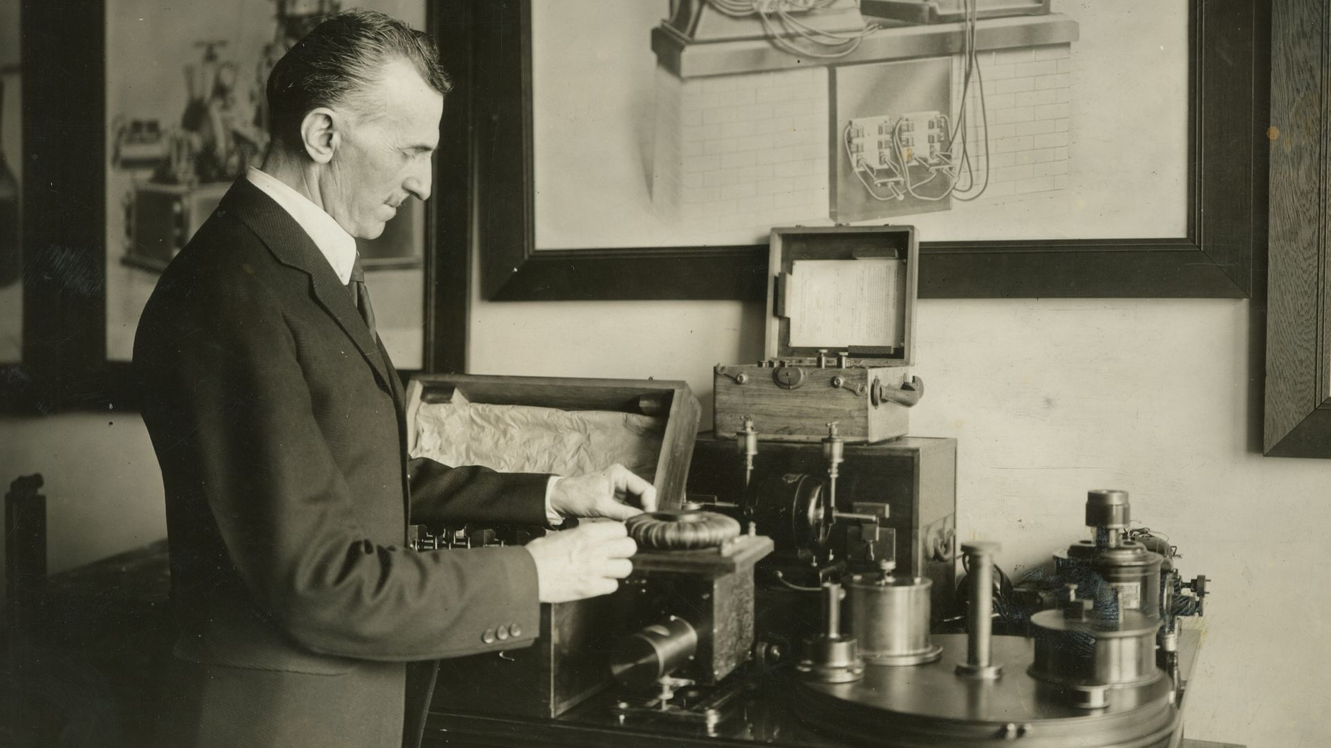 Nikola Tesla in his laboratory in New York. Behind Tesla are large framed photographs of his inventions.