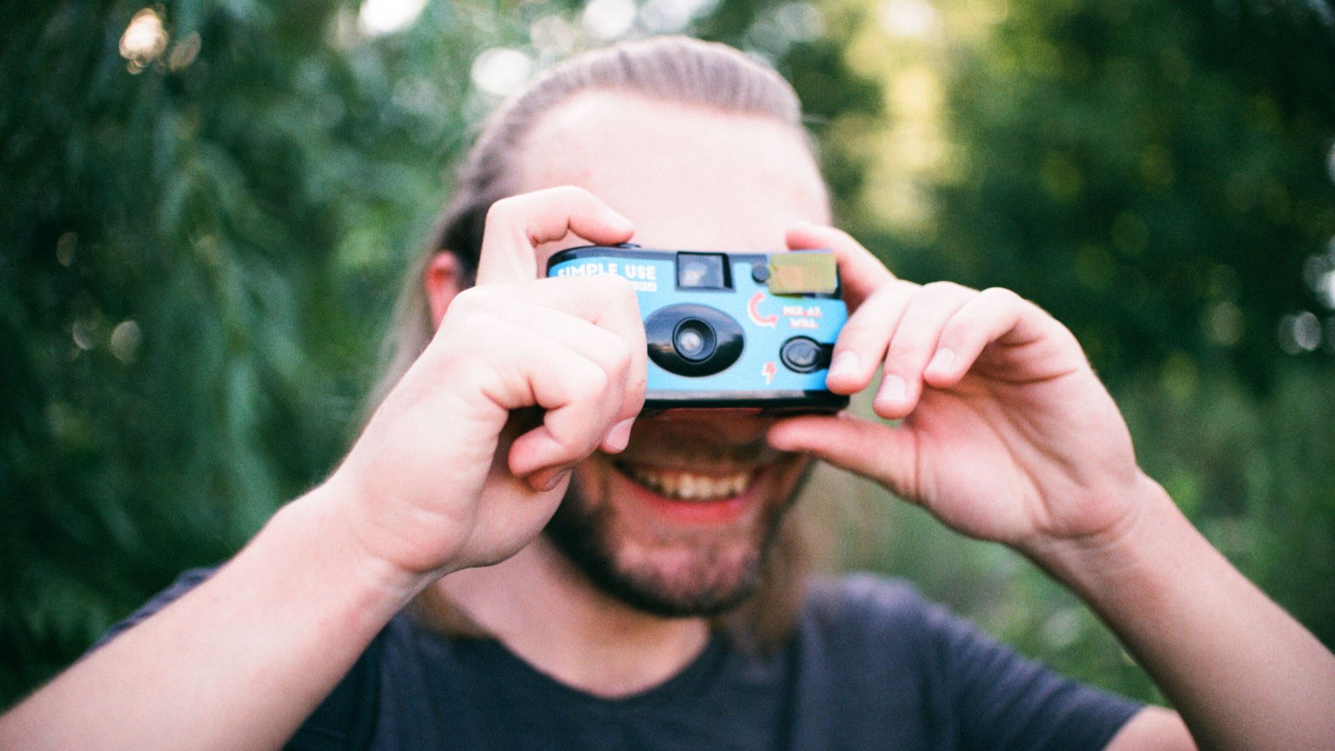 A smiling man taking photos with a camera in a lush outdoor setting, enjoying leisure time.