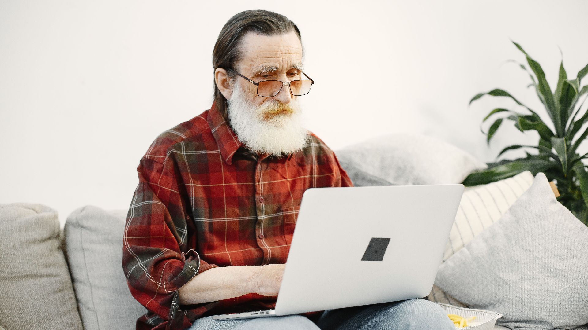 An elderly man using a laptop on a comfortable sofa in a home setting.
