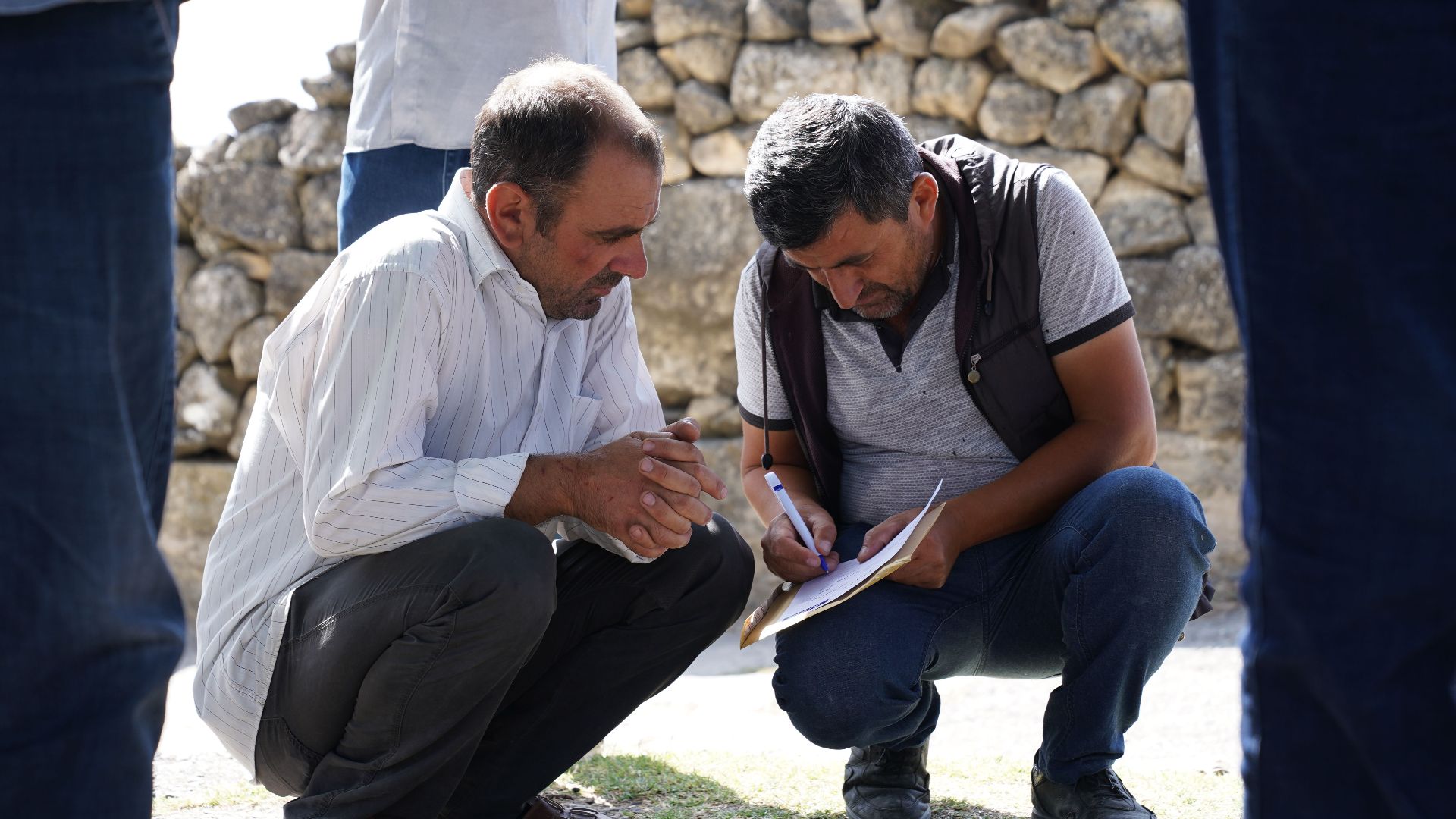 Two men squatting and analyzing paperwork outdoors in a sunny setting.