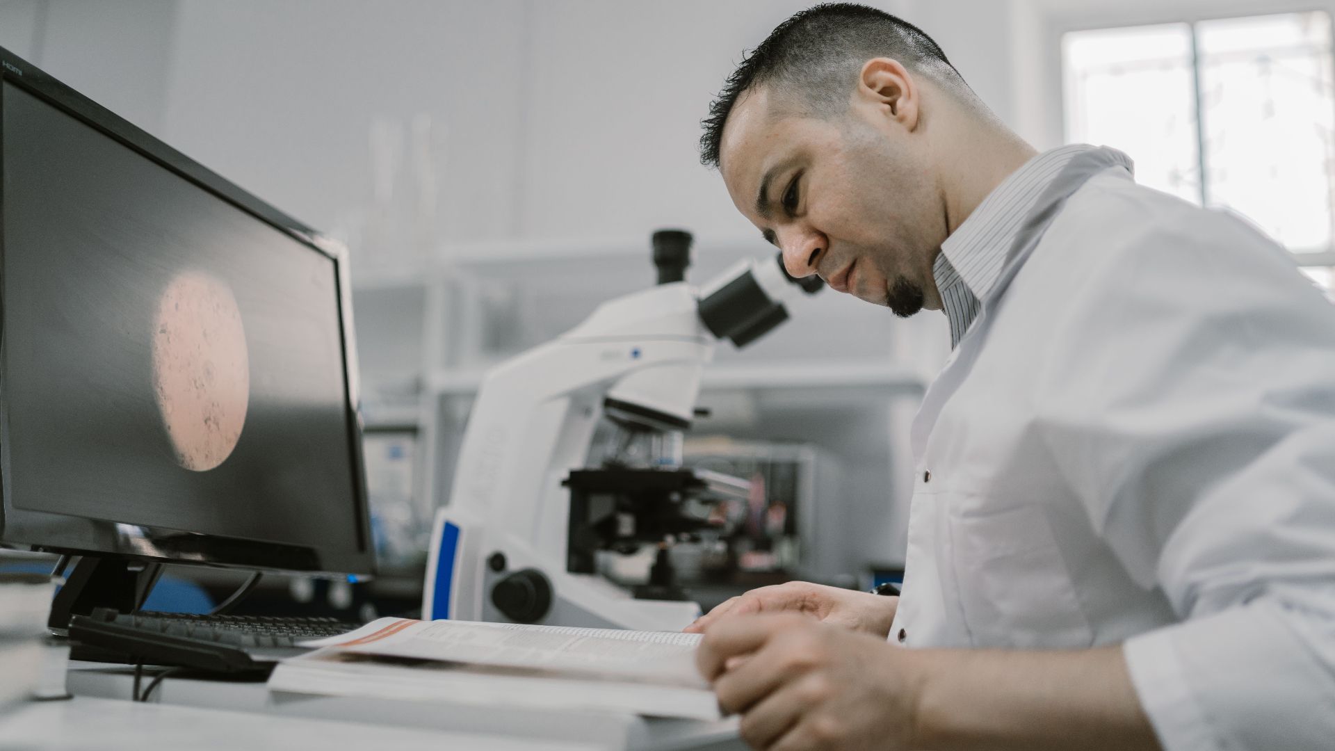 Asian man reading in a laboratory, surrounded by equipment and microscope, focused on work.