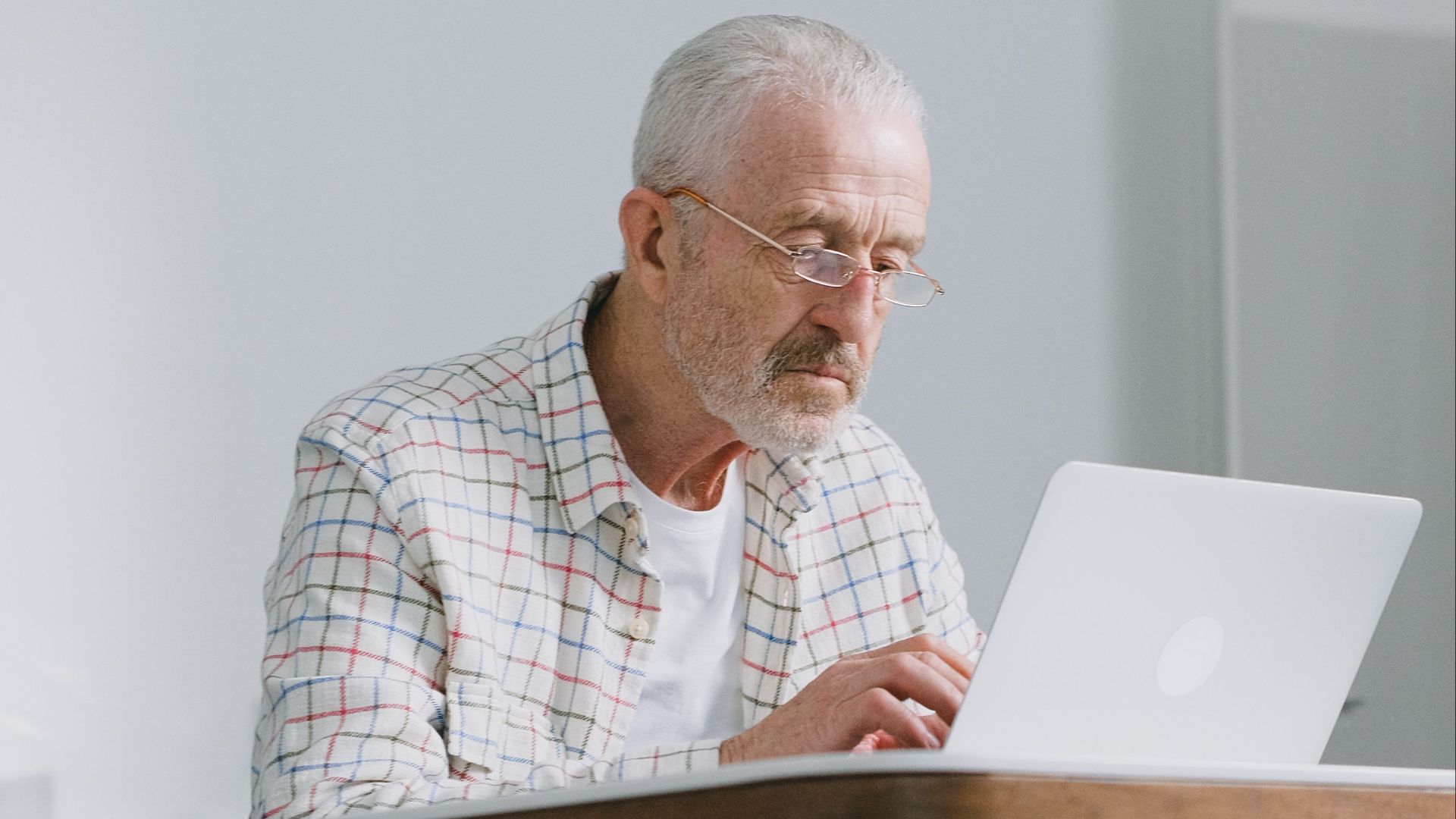 Elderly man focused on typing on laptop indoors. Natural lighting, modern ambiance.