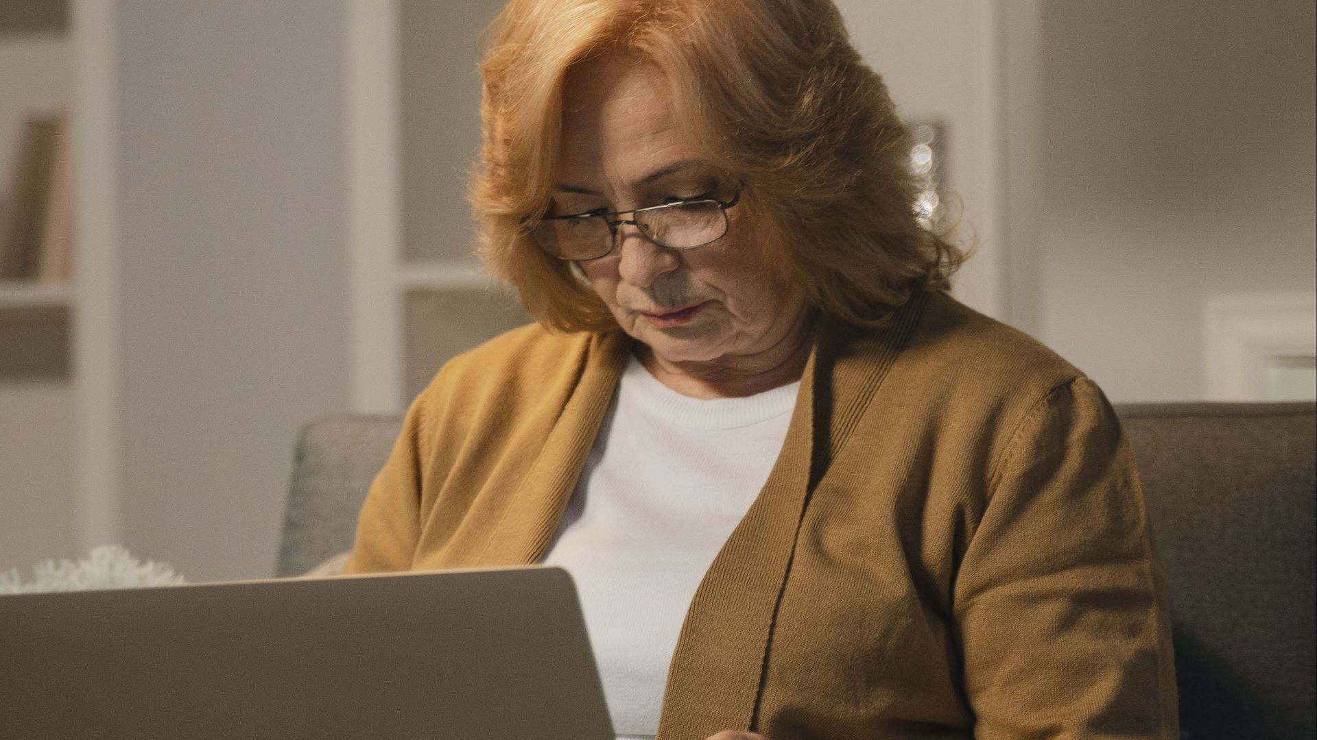 A senior woman with glasses sits on a sofa using a laptop in a cozy living room.
