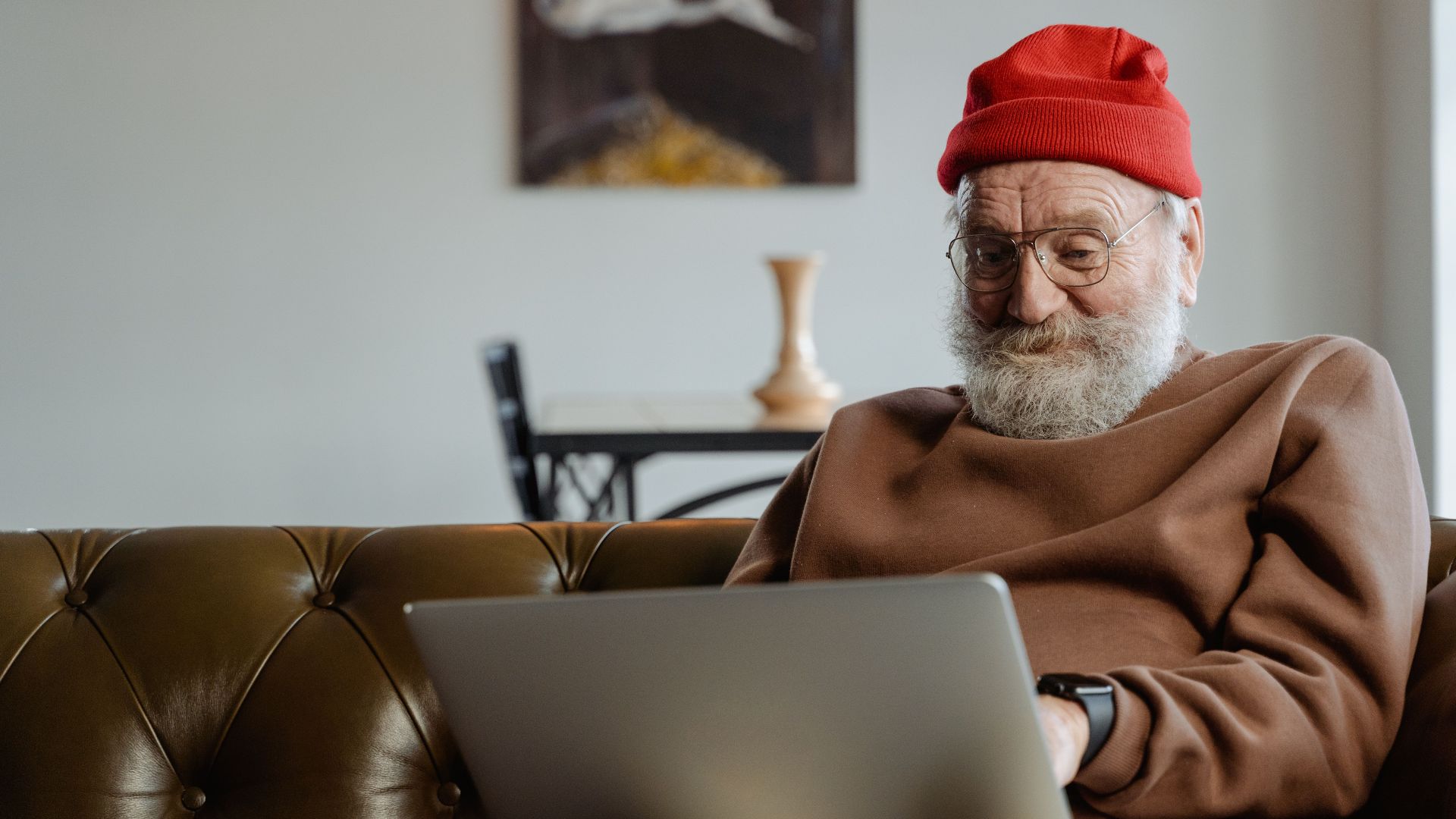 Senior man wearing a red hat and eyeglasses, using a laptop on a sofa indoors.