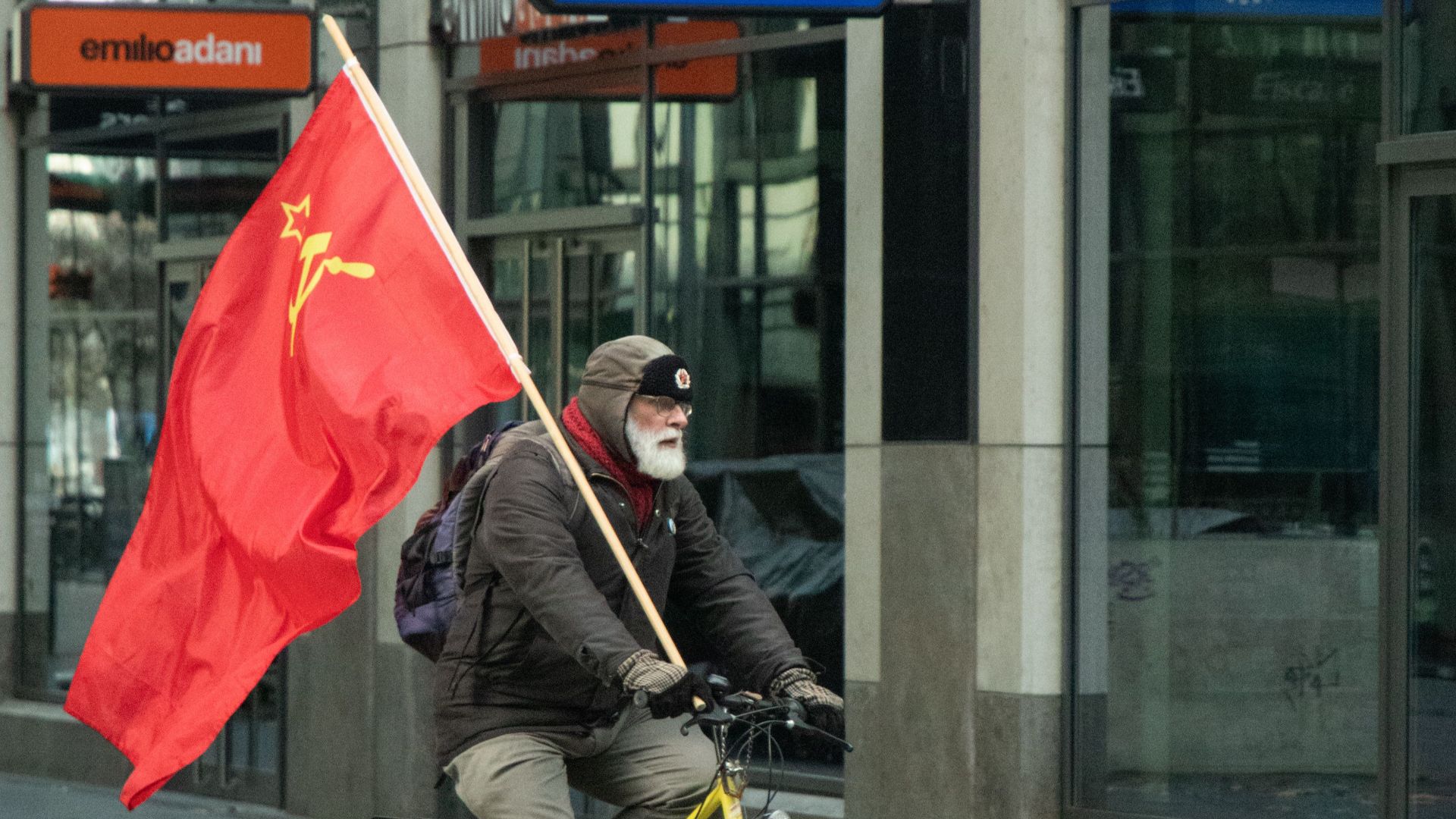 Senior adult riding bicycle with Soviet flag on busy urban street, evoking historical themes.