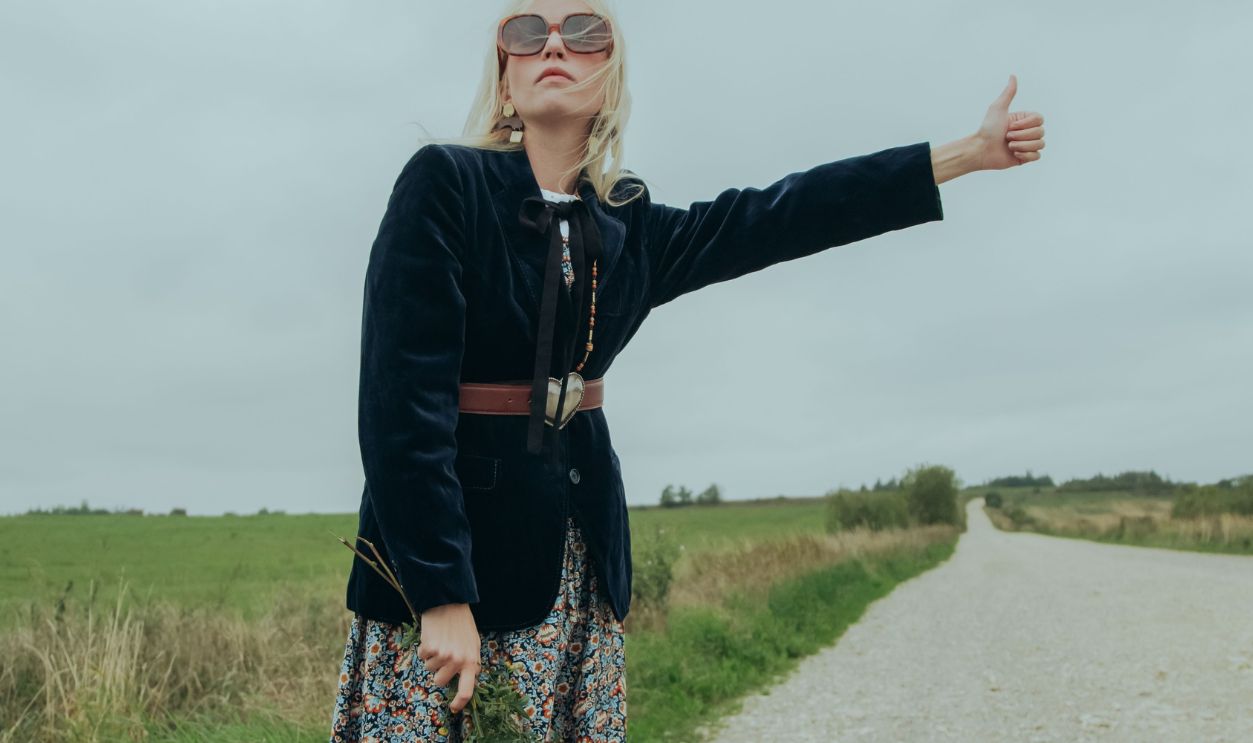 Stylish woman in floral skirt hitchhiking on a rural road with a thumb up, wearing sunglasses
