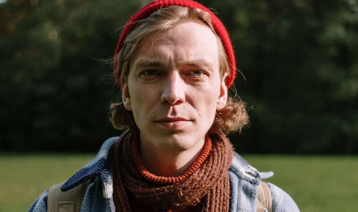A man in a plaid jacket and knitted scarf stands in a sunny park, surrounded by trees