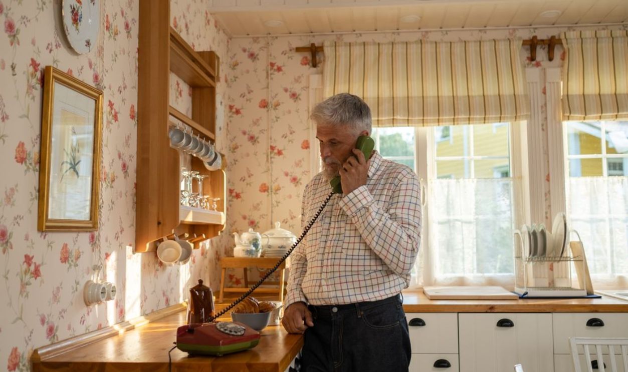 Senior man talks on a vintage rotary phone in a retro-styled kitchen with floral wallpaper and classic decor