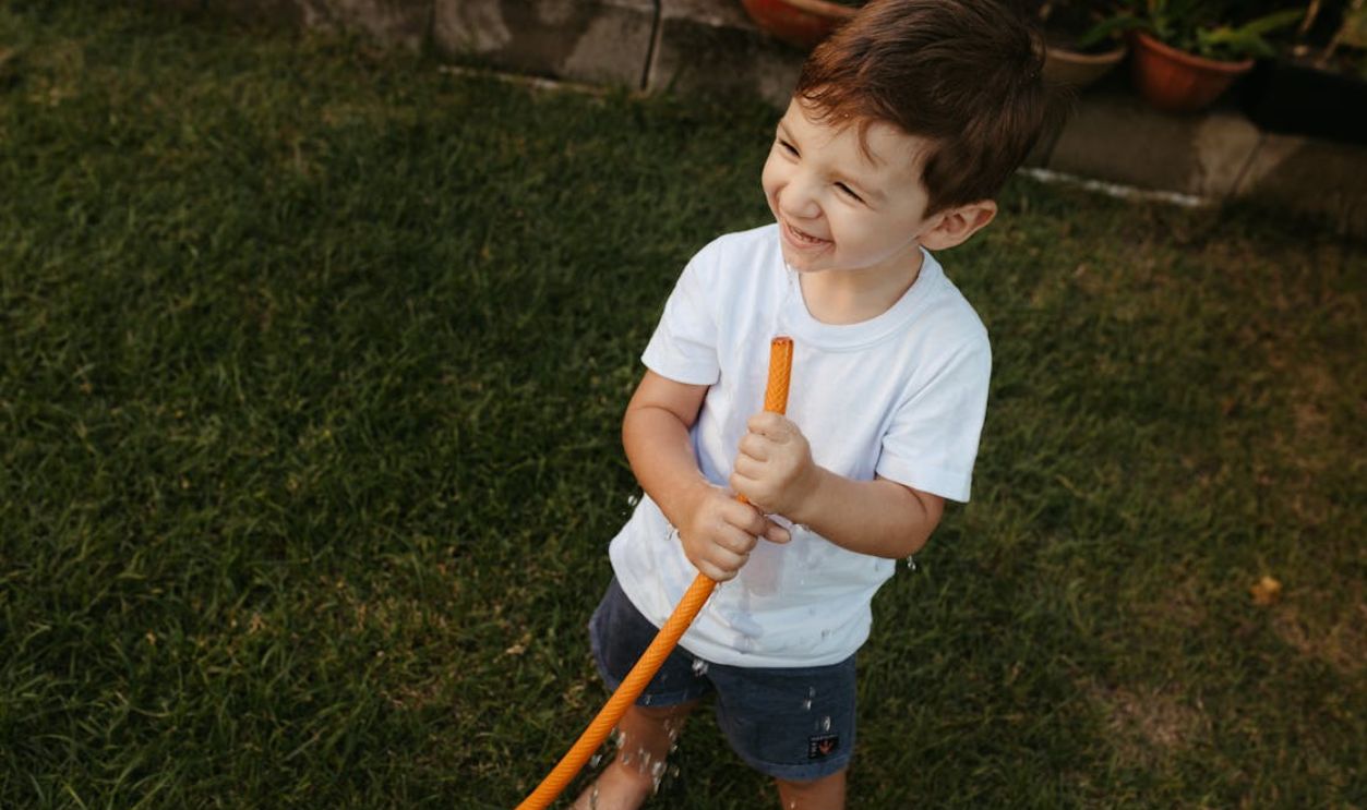 A smiling child enjoys playfully sprinkling water with a hose in a sunny garden
