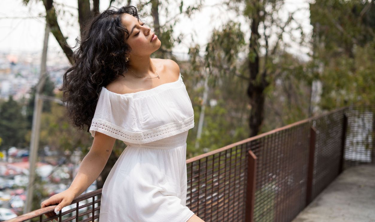 A young woman in a white dress leans on a railing in an outdoor setting, enjoying nature.
